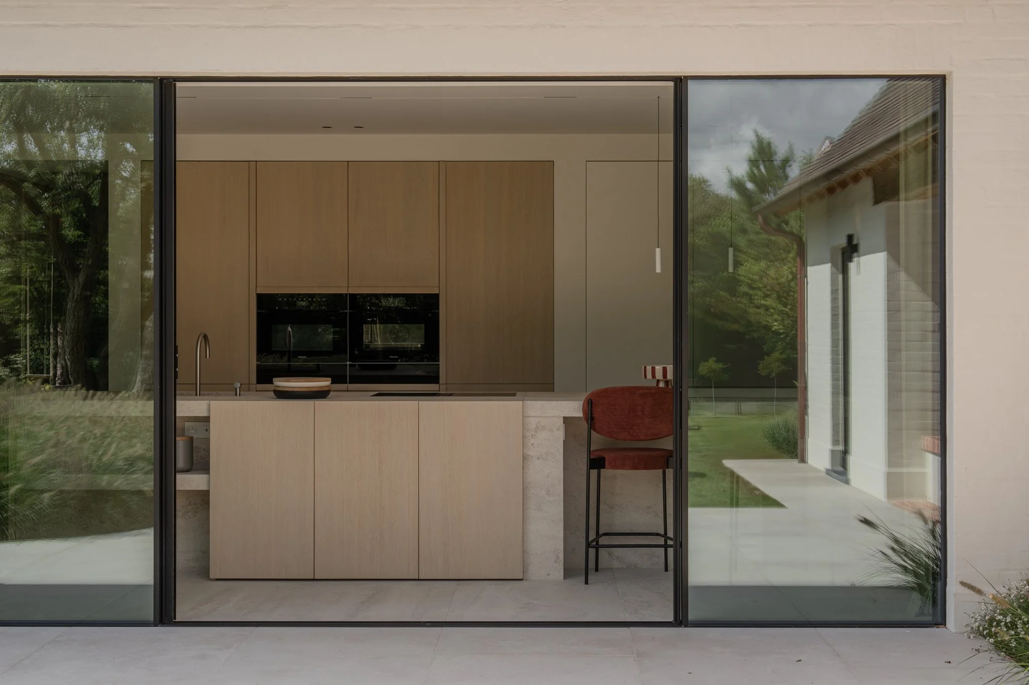 Modern kitchen with wooden cabinets, beige island, and a red chair outside the glass sliding door, with a green outdoor area visible.