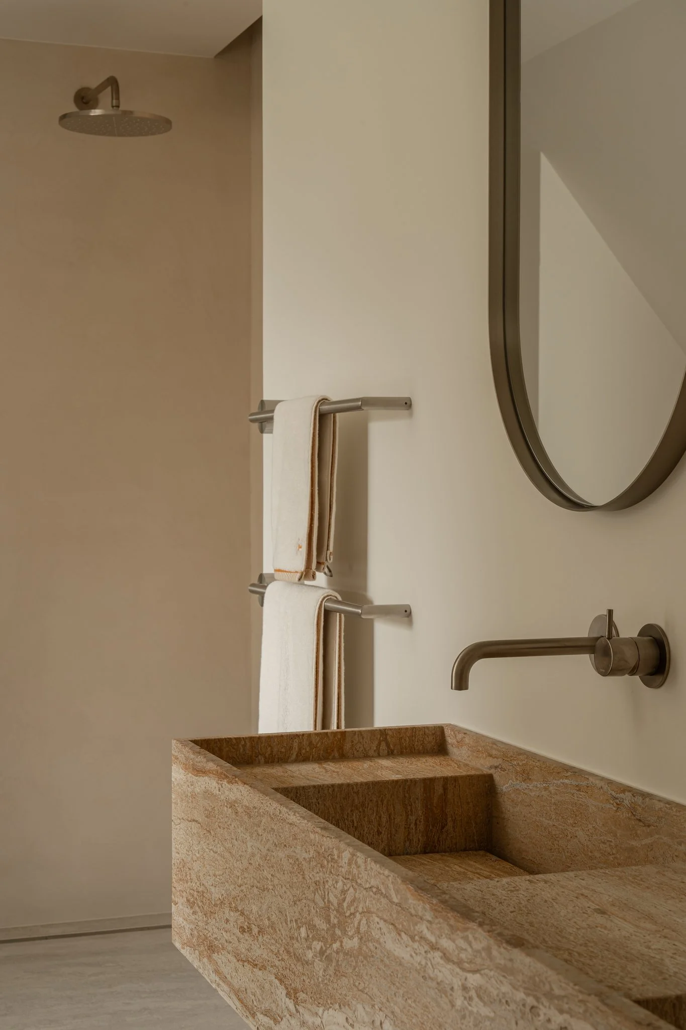 A bathroom with a beige stone sink, a wall-mounted metallic faucet, a large oval mirror, a towel rack with two white towels, and a showerhead in the background.