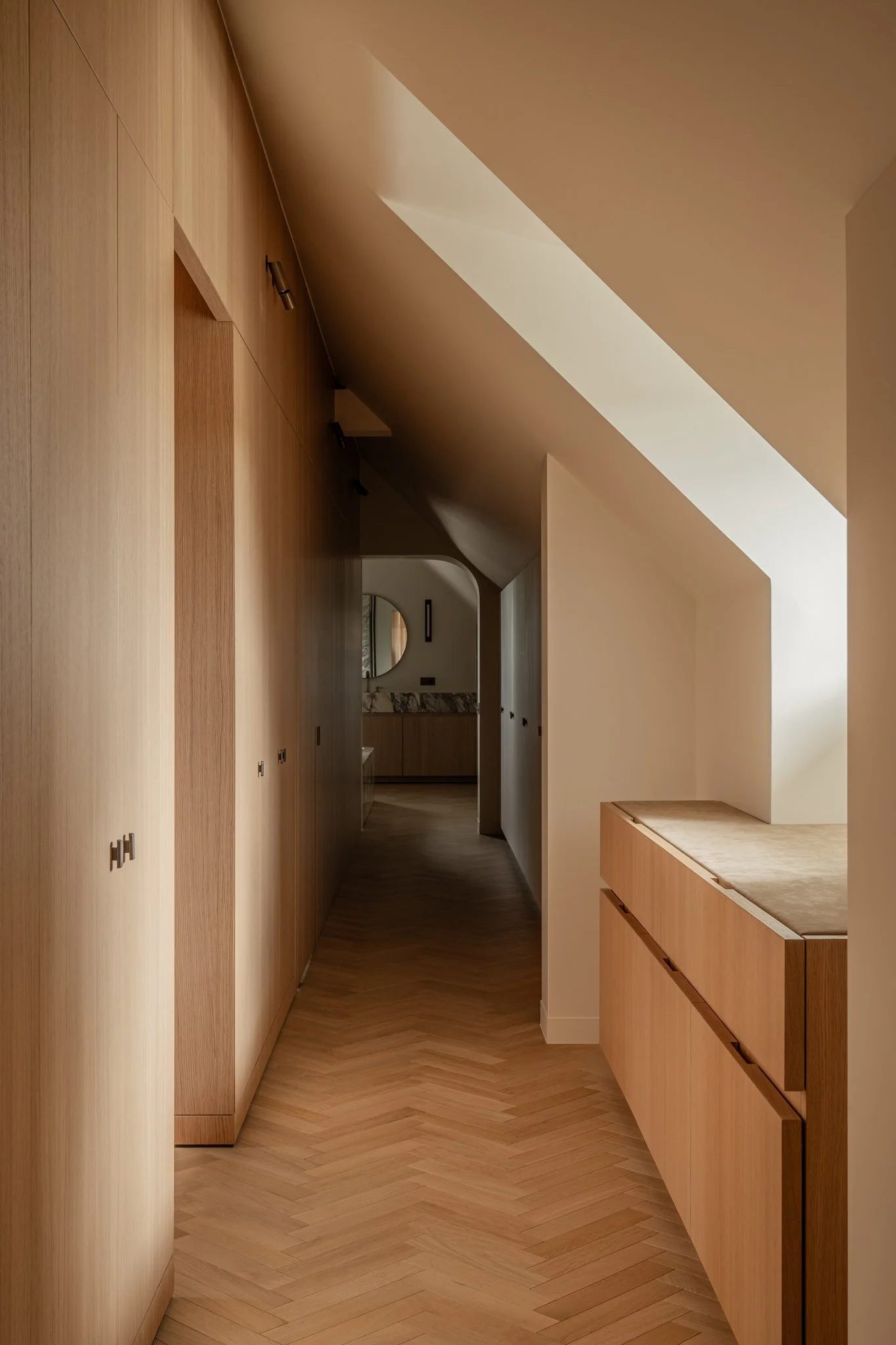 A minimalist hallway with wooden cabinets and a sloped ceiling with a skylight, leading to a bathroom with a marble countertop and oval mirror.