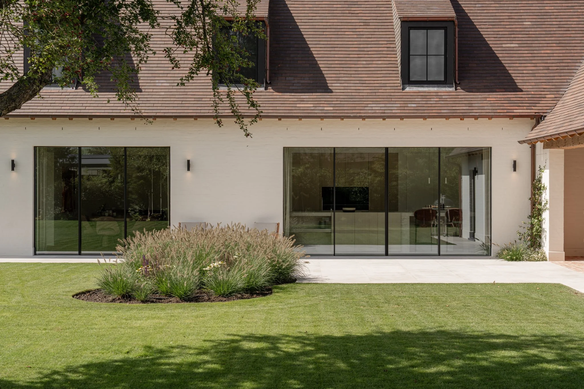 A modern house with a white exterior wall, large glass sliding doors, a tiled patio, a well-maintained lawn, and a flower bed with ornamental grasses and small flowers. The house has a brown shingle roof and small black wall-mounted lights.