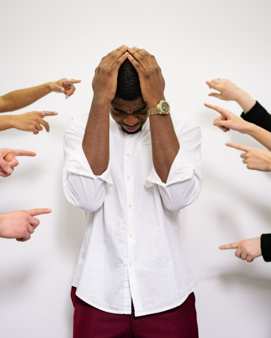 Man in white shirt surrounded by pointing fingers, holding head in distress