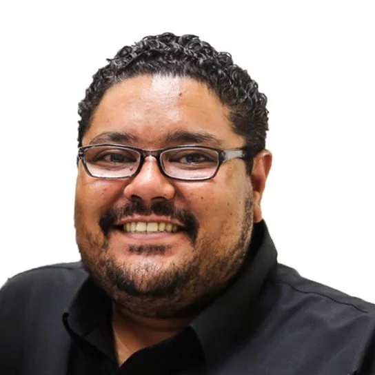 Close-up photo of a smiling man with curly hair, glasses, a beard, wearing a black shirt, against a plain white background.