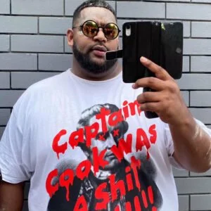 Man taking a selfie in front of a gray brick wall, wearing sunglasses and a white T-shirt with red and black graphic text.