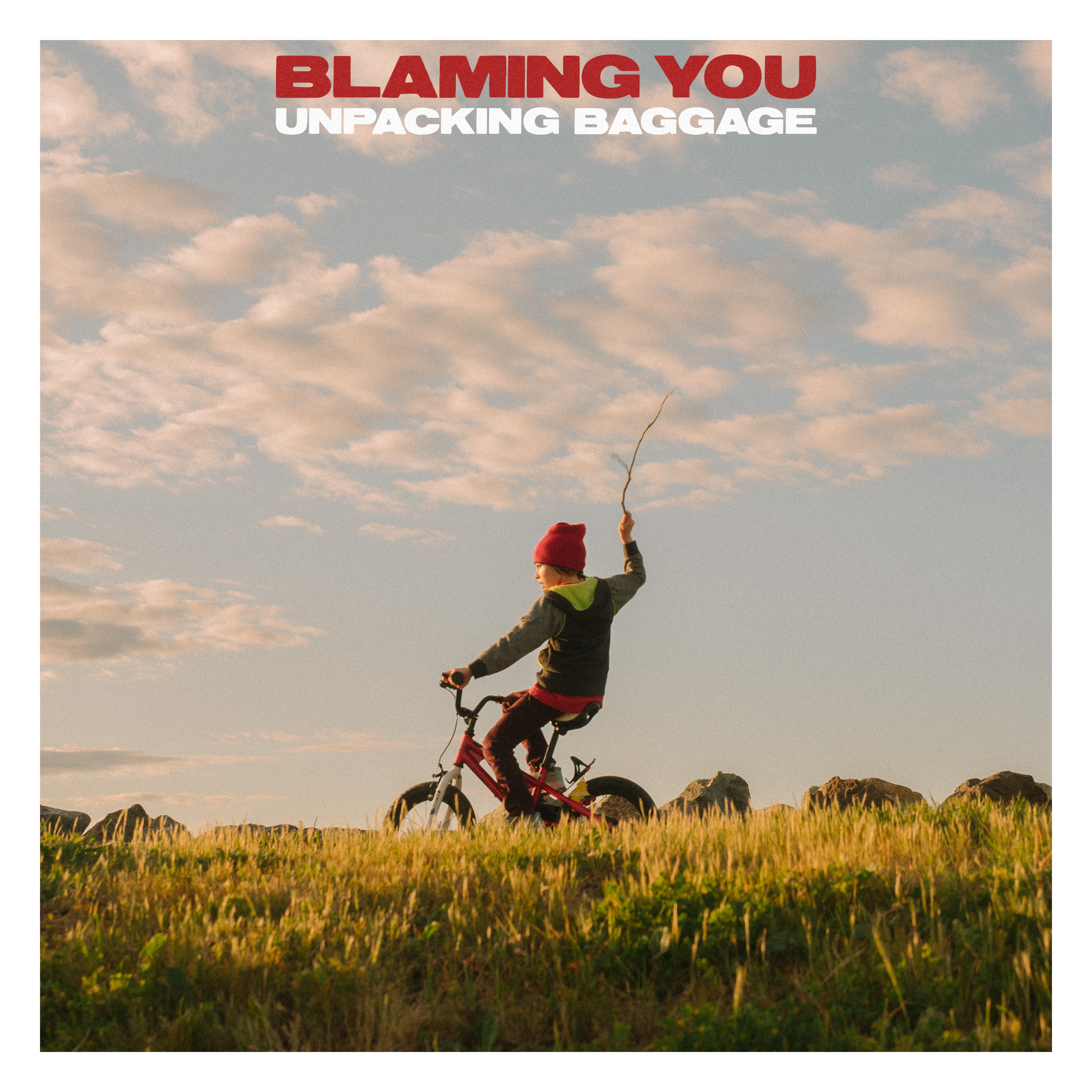 Child riding a red bicycle across a grassy field at sunset, wearing a red hat and a dark hoodie with green lining.