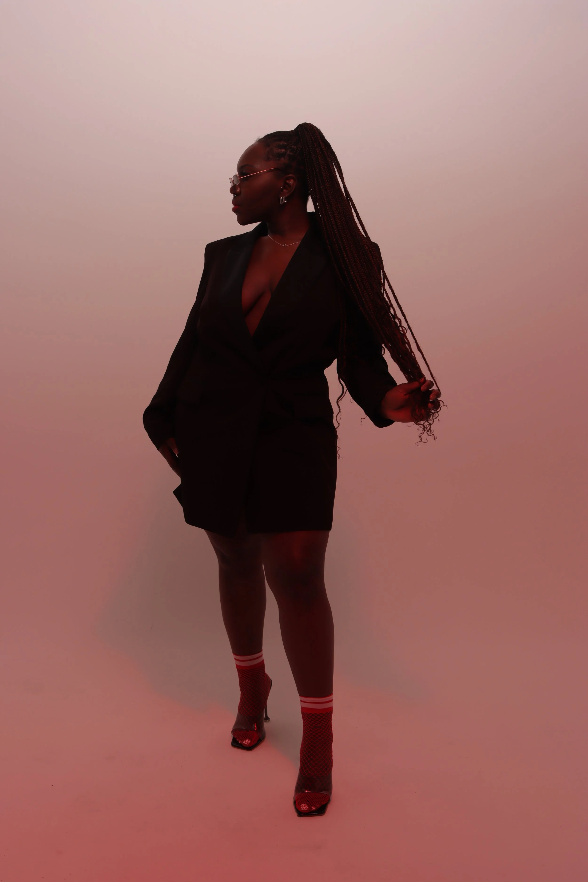A woman with long braids wearing glasses, a black blazer dress, red fishnet socks, and high heels, standing against a plain wall with a pinkish hue.