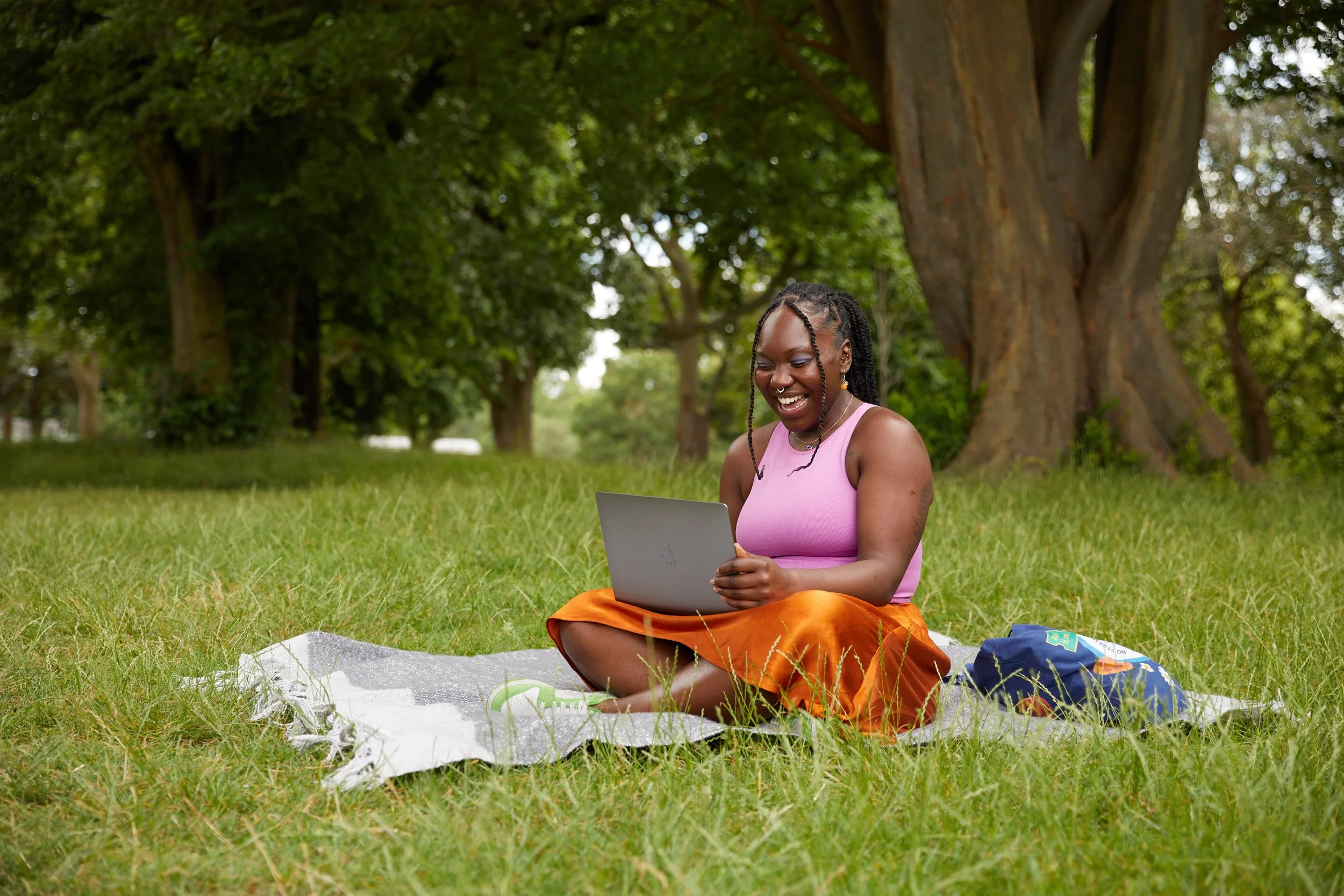 A young woman sitting on a blanket in a park, smiling and looking at her laptop, with large trees and green grass around her.