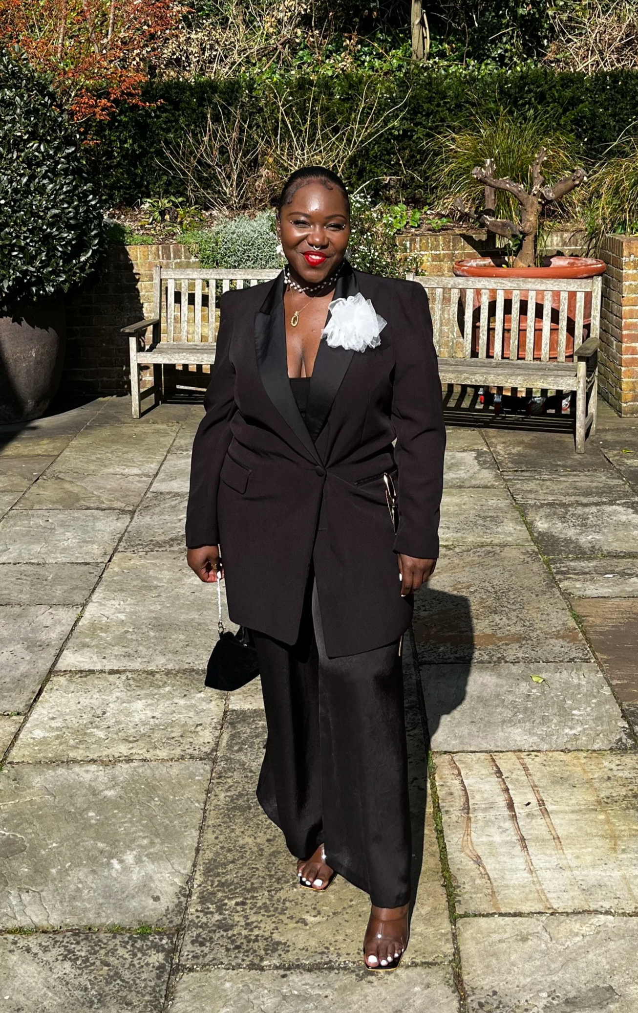 Tamara standing outdoors on a stone patio, dressed in a black blazer, black pants, and open-toed heels. She has jewelry including a necklace and earrings, and a white floral accessory on her blazer. Behind her are benches, a garden, and shrubs.