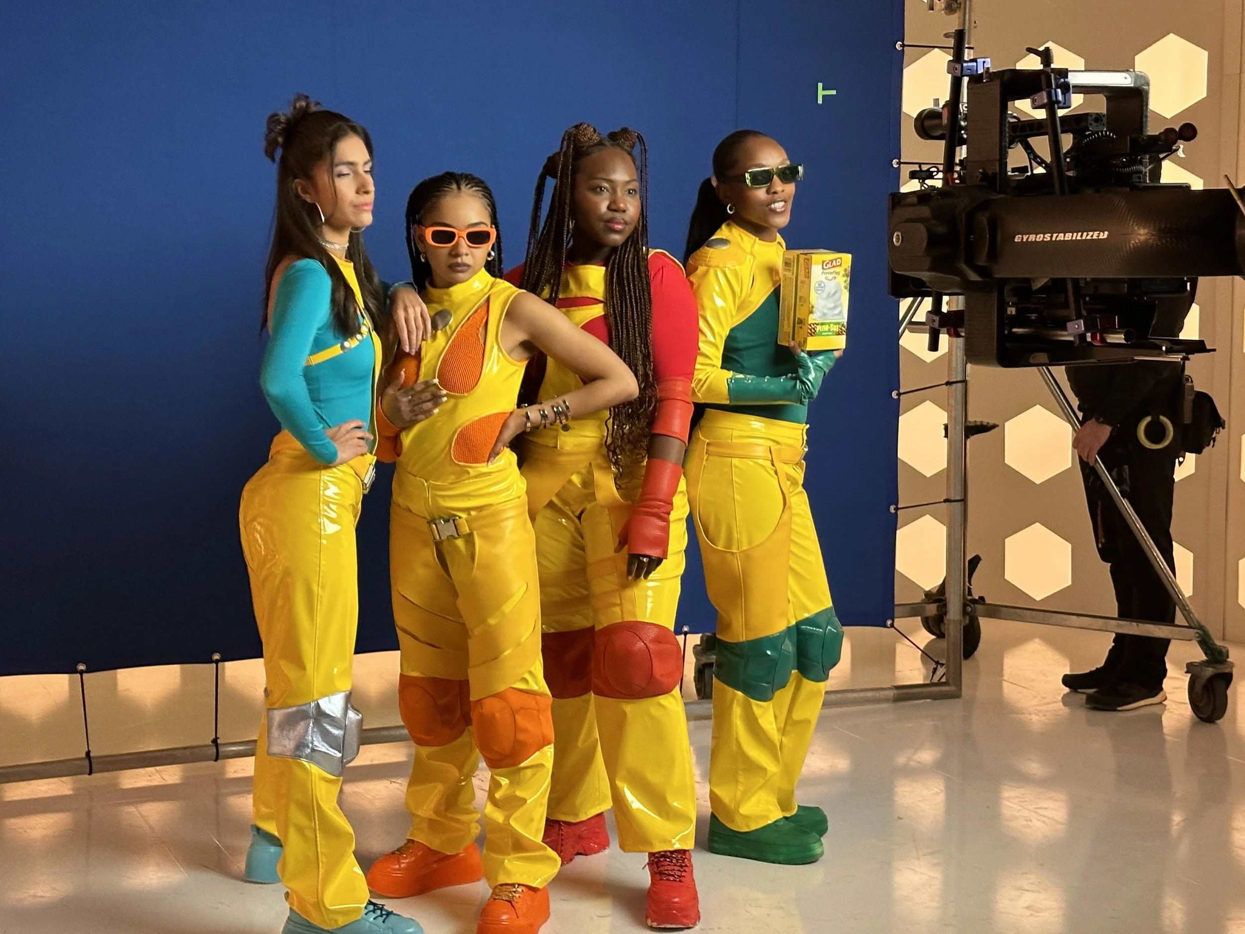 Four women dressed in colorful, futuristic outfits and sunglasses, standing in front of a blue screen on a studio set, with a camera and crew member in the background.