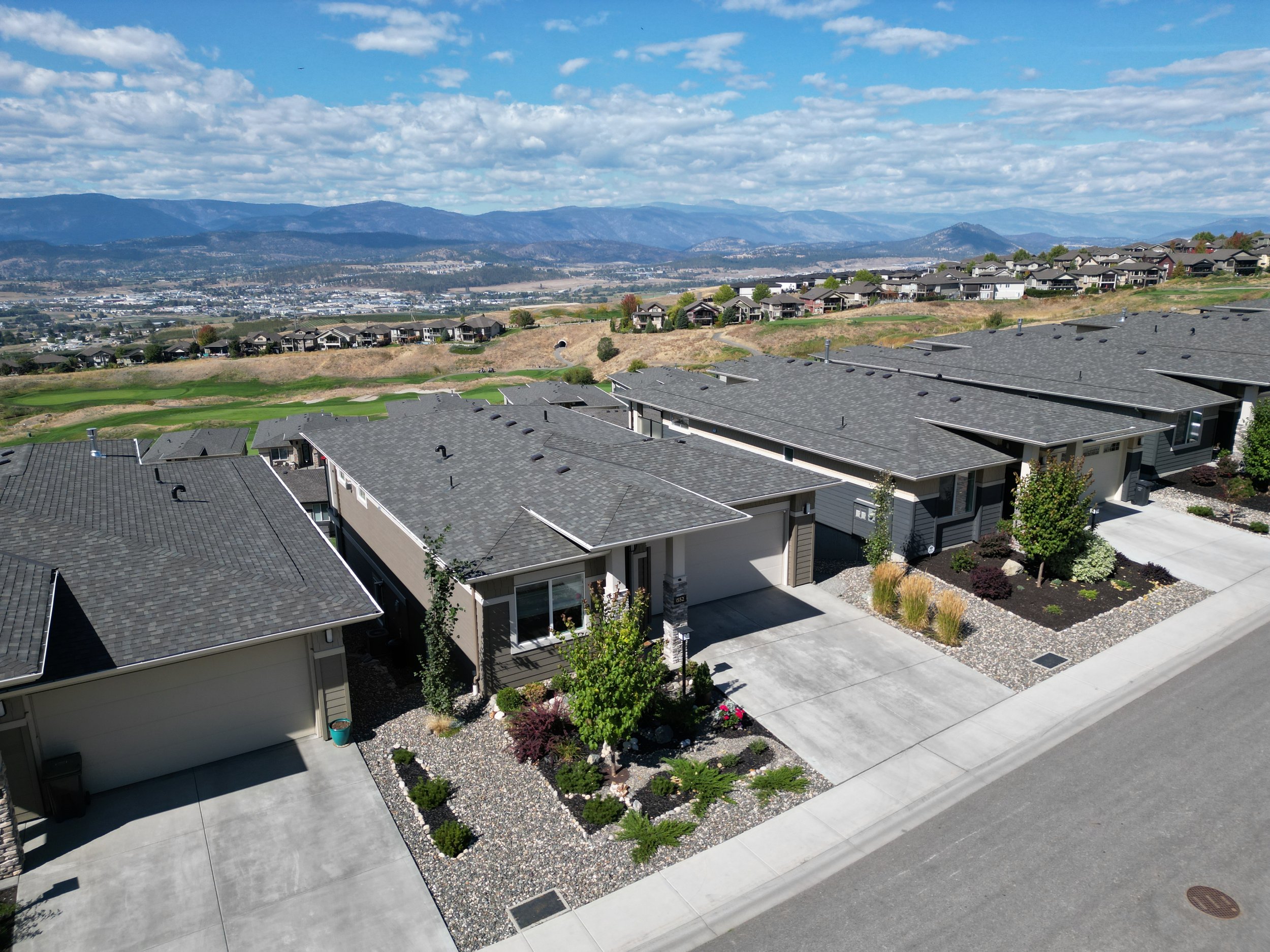 Aerial view of a suburban neighborhood with row houses, landscaped front yards, and concrete driveways. Background shows rolling hills and mountains under a partly cloudy sky.