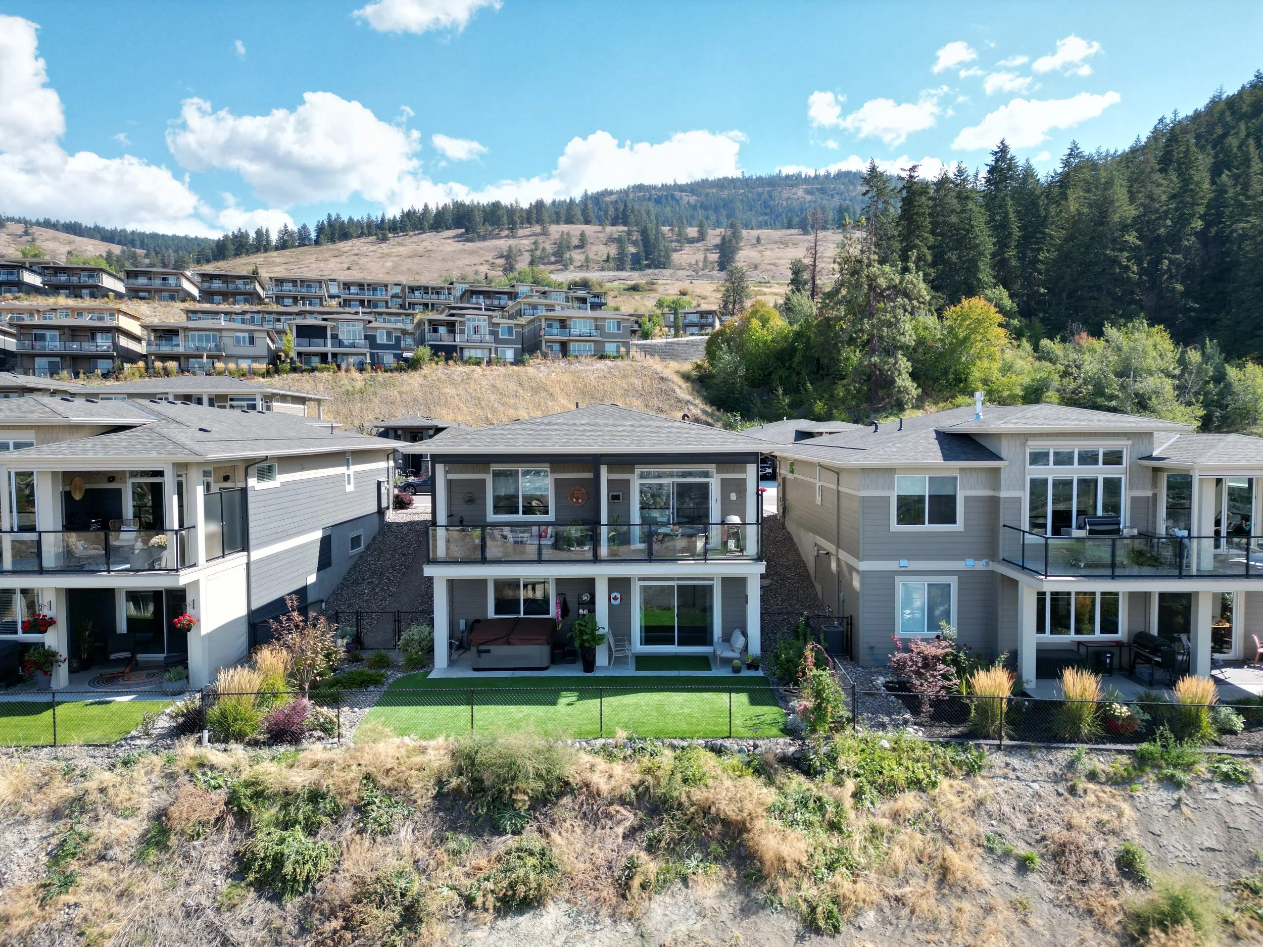 View of modern duplex-style houses with balconies, situated on a hill with landscaped lawns and greenery. Background features additional homes on elevated terrain and a forested area under a clear blue sky with clouds.