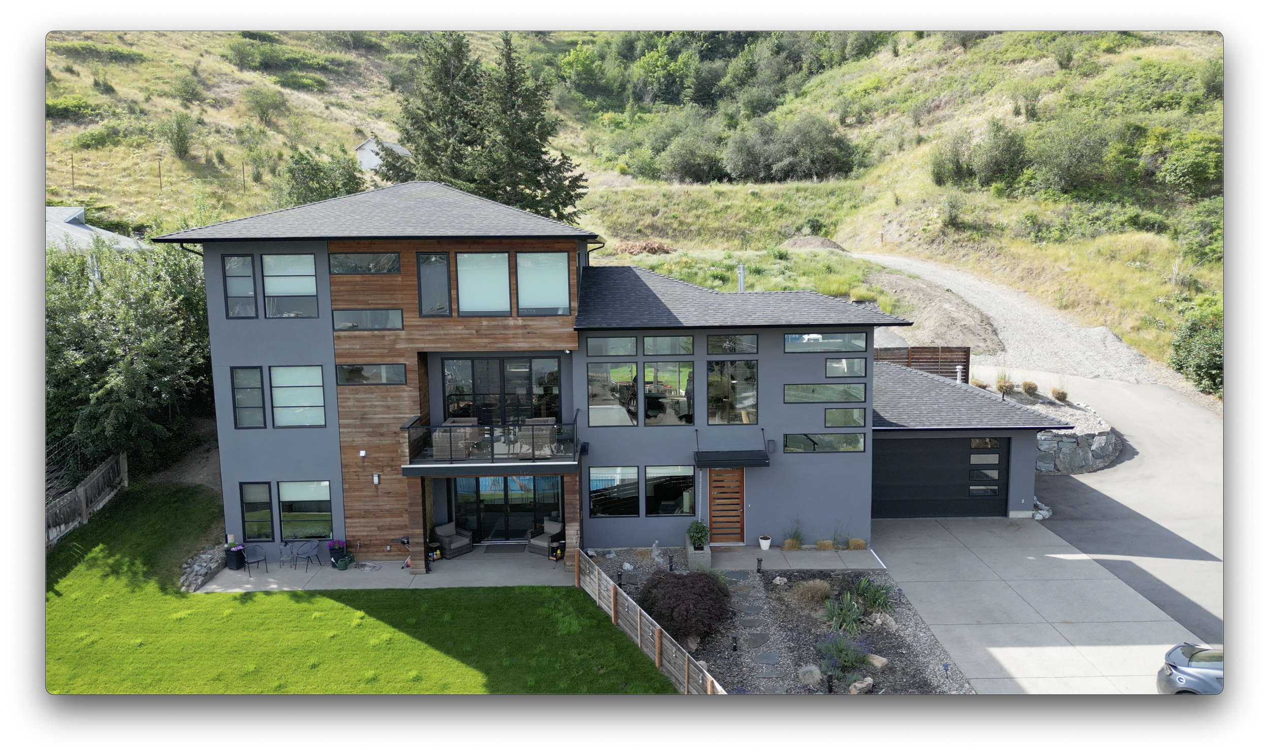 Modern two-story house with wooden and gray siding, large windows, a balcony, and a well-maintained lawn, set against a hillside backdrop.
