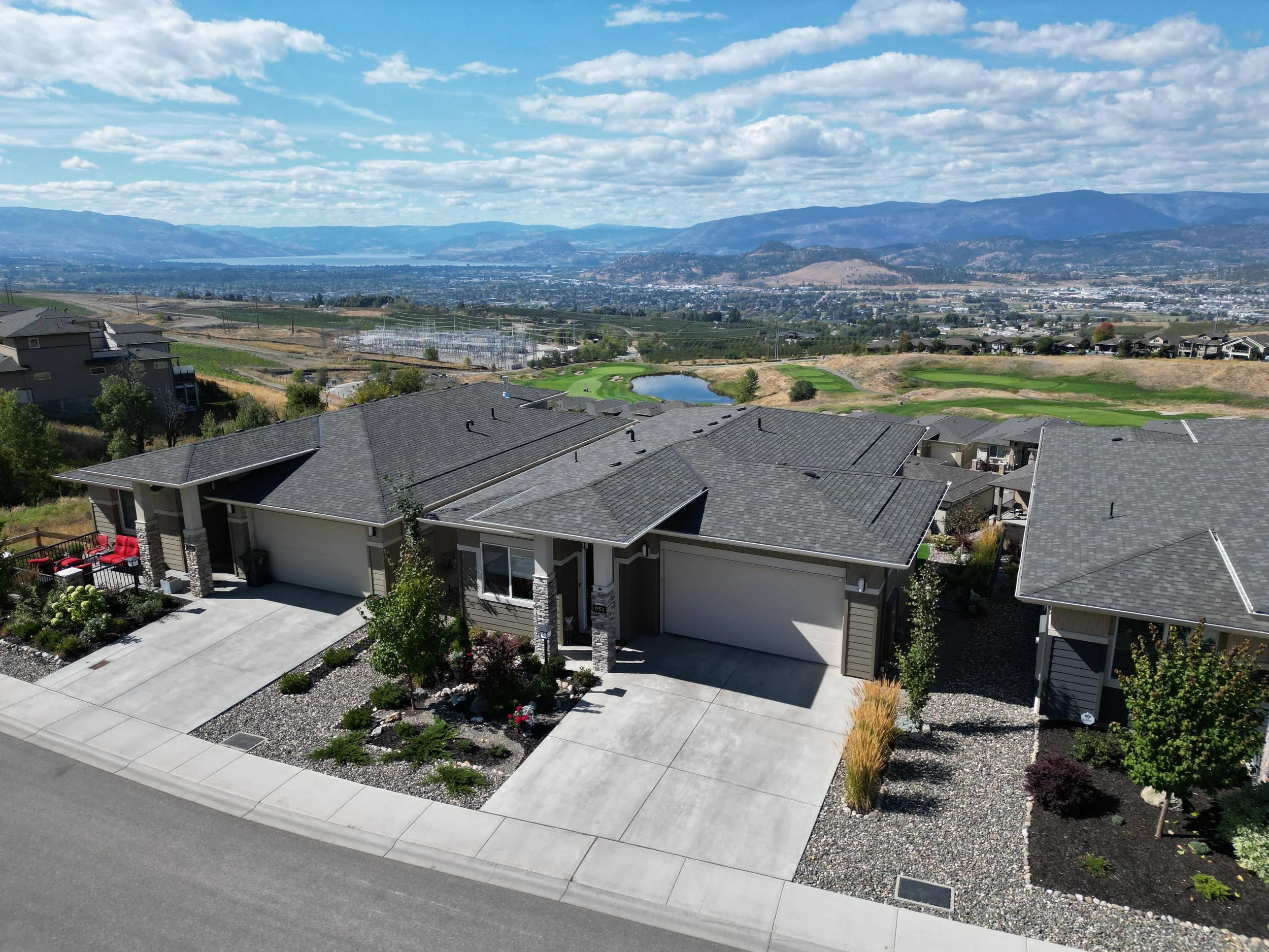 Aerial view of suburban houses with gray roofs, a paved street, landscaped yards with shrubs, and a scenic background of mountains, water, and golf course.