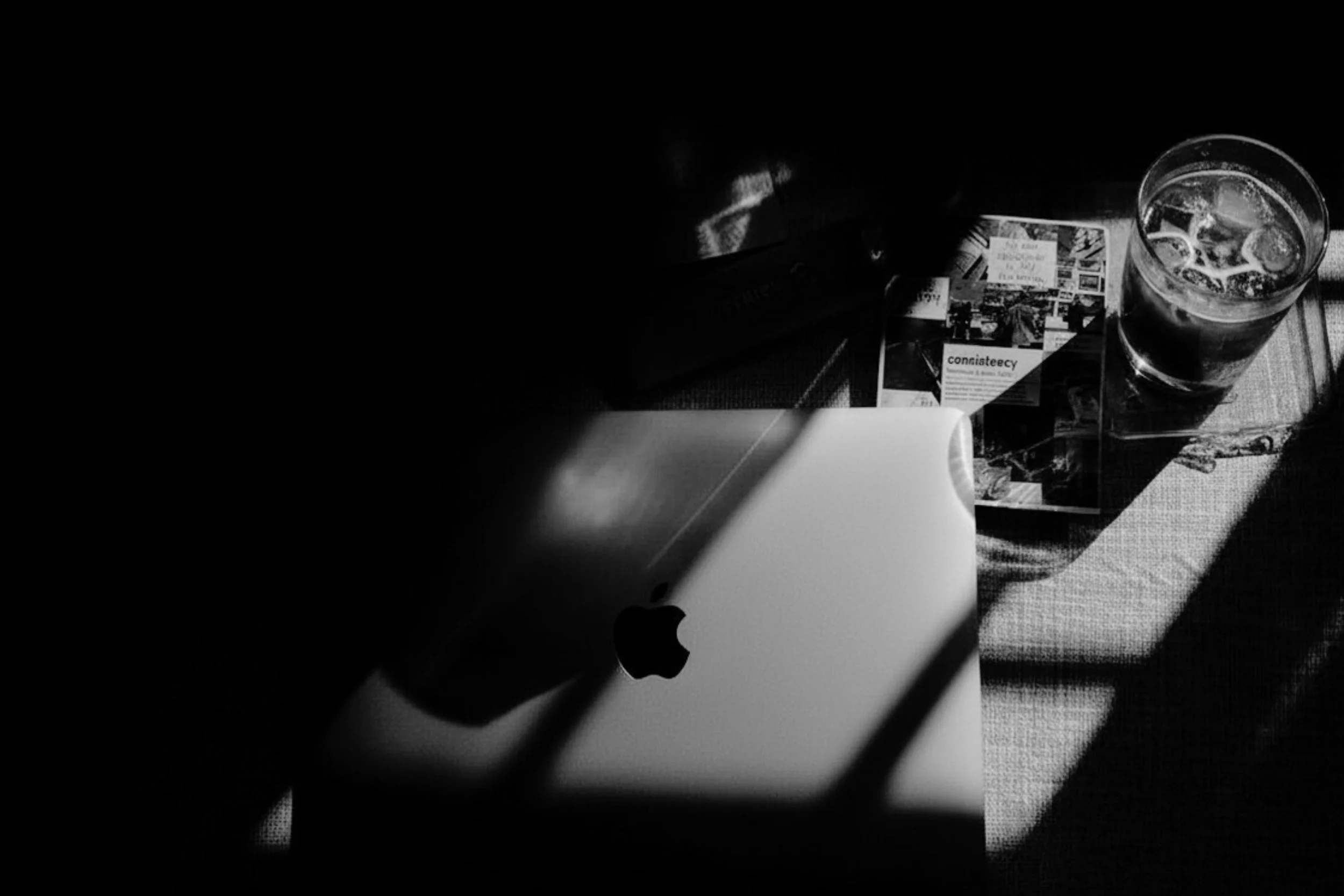 Moody black and white photo of a MacBook laptop on a desk beside an iced drink and scattered papers