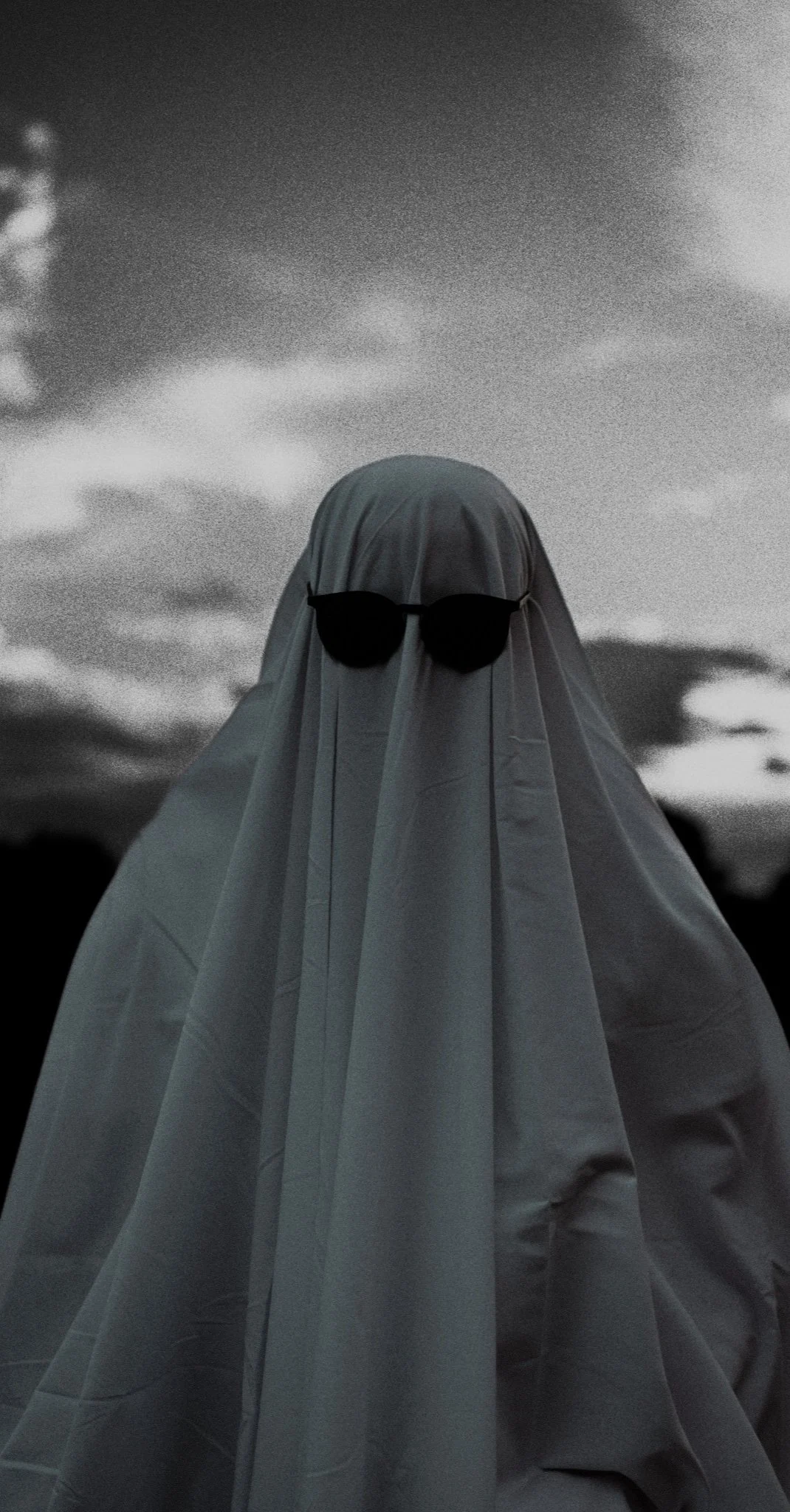 Grainy black and white photo of a figure draped in a sheet wearing sunglasses against a cloudy sky