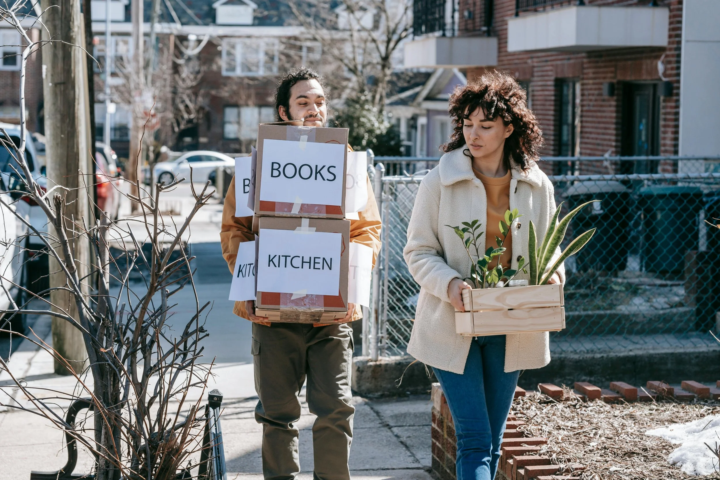 Two people walk down a city street—one carrying stacked boxes, the other holding a plant—capturing the emotional weight and uncertainty of major life changes and transitions.