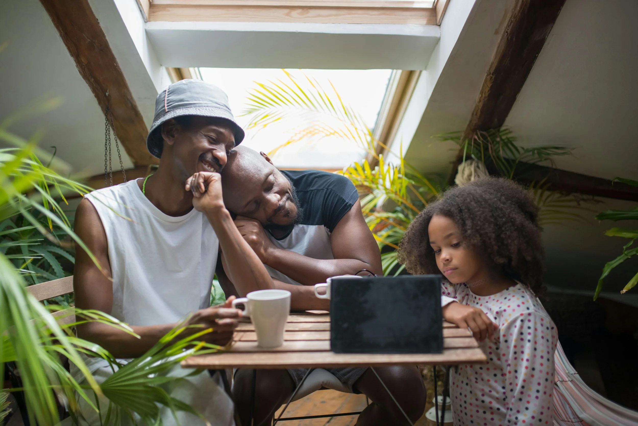 A family sits together at a table—two adults and a child. The child looks down, disengaged, while one adult leans into the other—reflecting emotional disconnection and complex family dynamics.