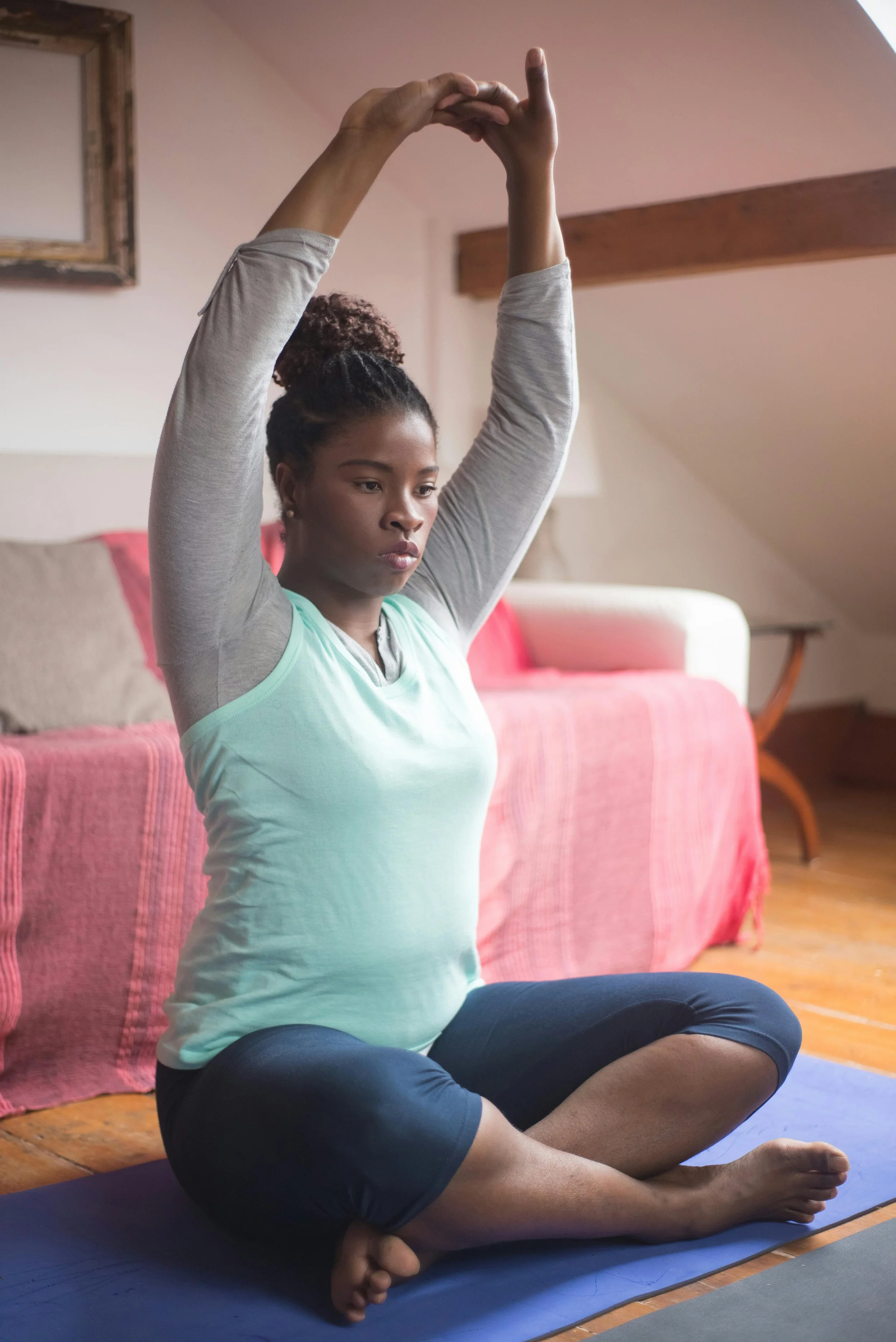 A woman sits in front of a couch, gently stretching her arms overhead with a relaxed, peaceful expression—capturing a moment of embodied movement and nervous system regulation.