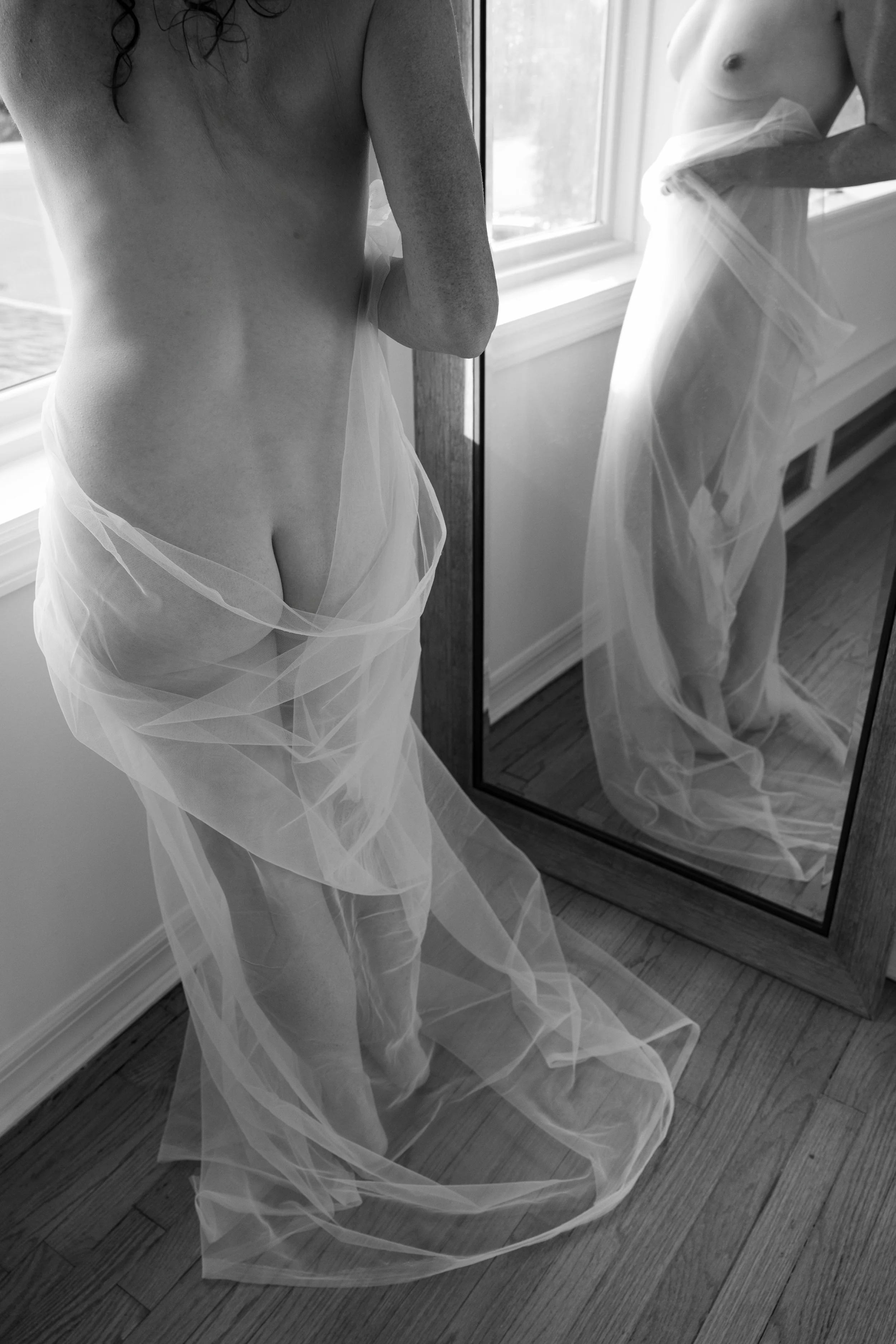 Nude boudoir portrait of a woman facing a mirror, her body wrapped in sheer fabric by Eye Candy Boudoir. 