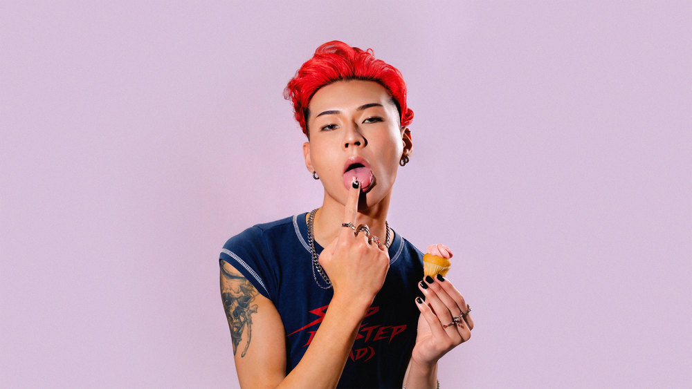 Kevin Silvester with red hair holding a pink-frosted cupcake against a lilac backdrop