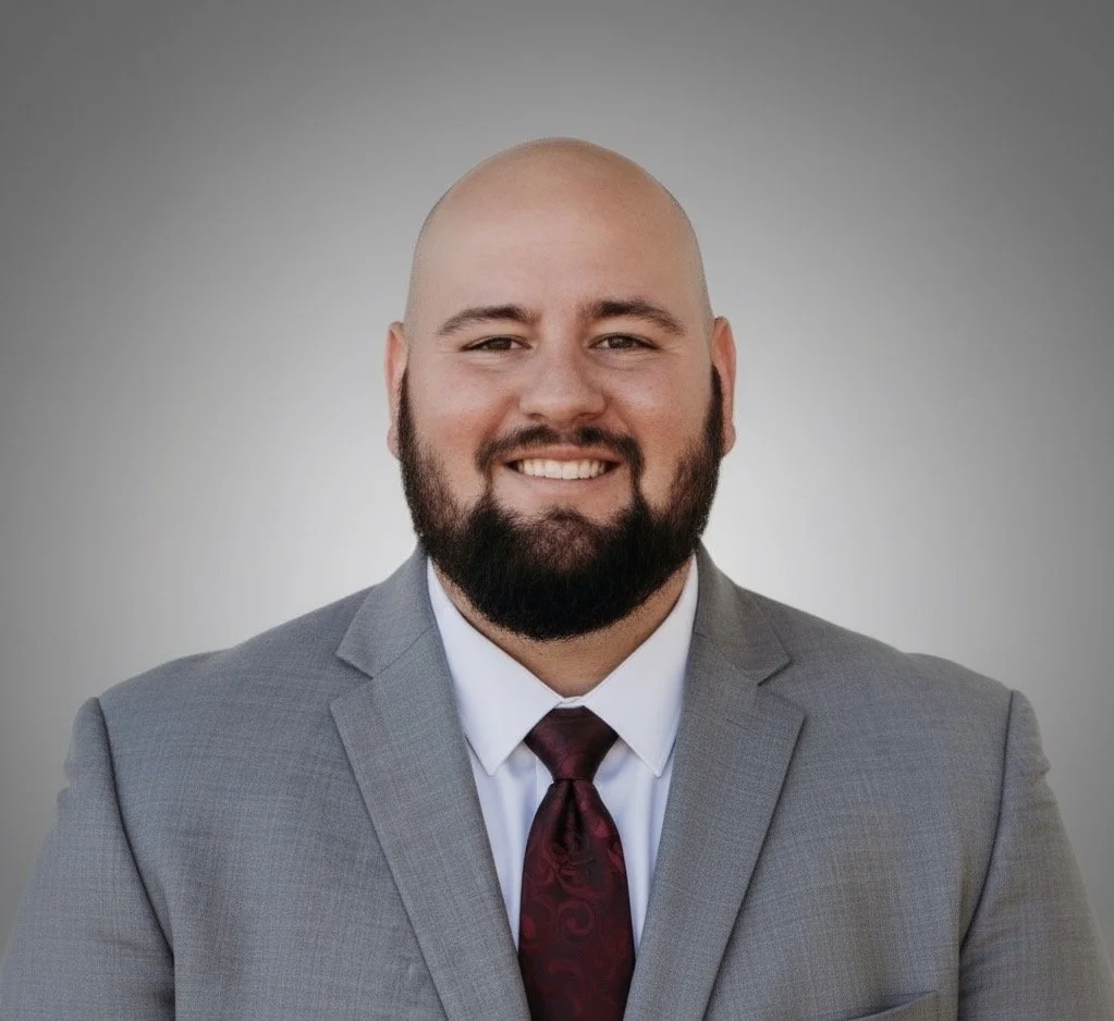 A man with a bald head and beard, wearing a gray suit, white shirt, and maroon tie, smiling in front of a plain gray background.