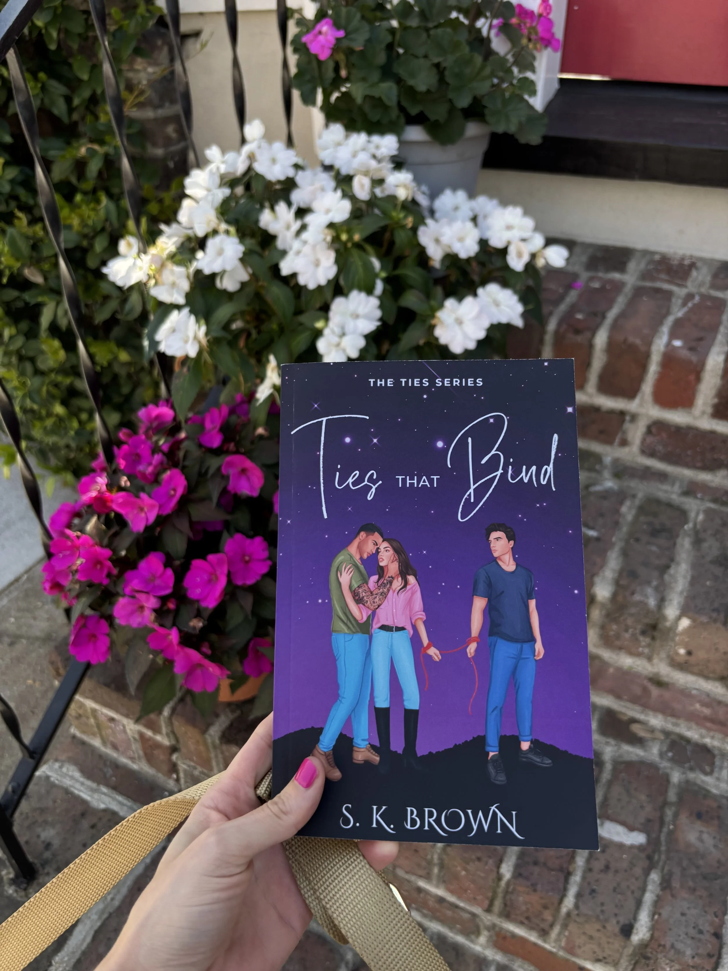 A person holding a book titled "Ties That Bind" by S. K. Brown in front of pink and white flowers outdoors. The background includes a fence, brick, and a window with pink flowers in a white pot.