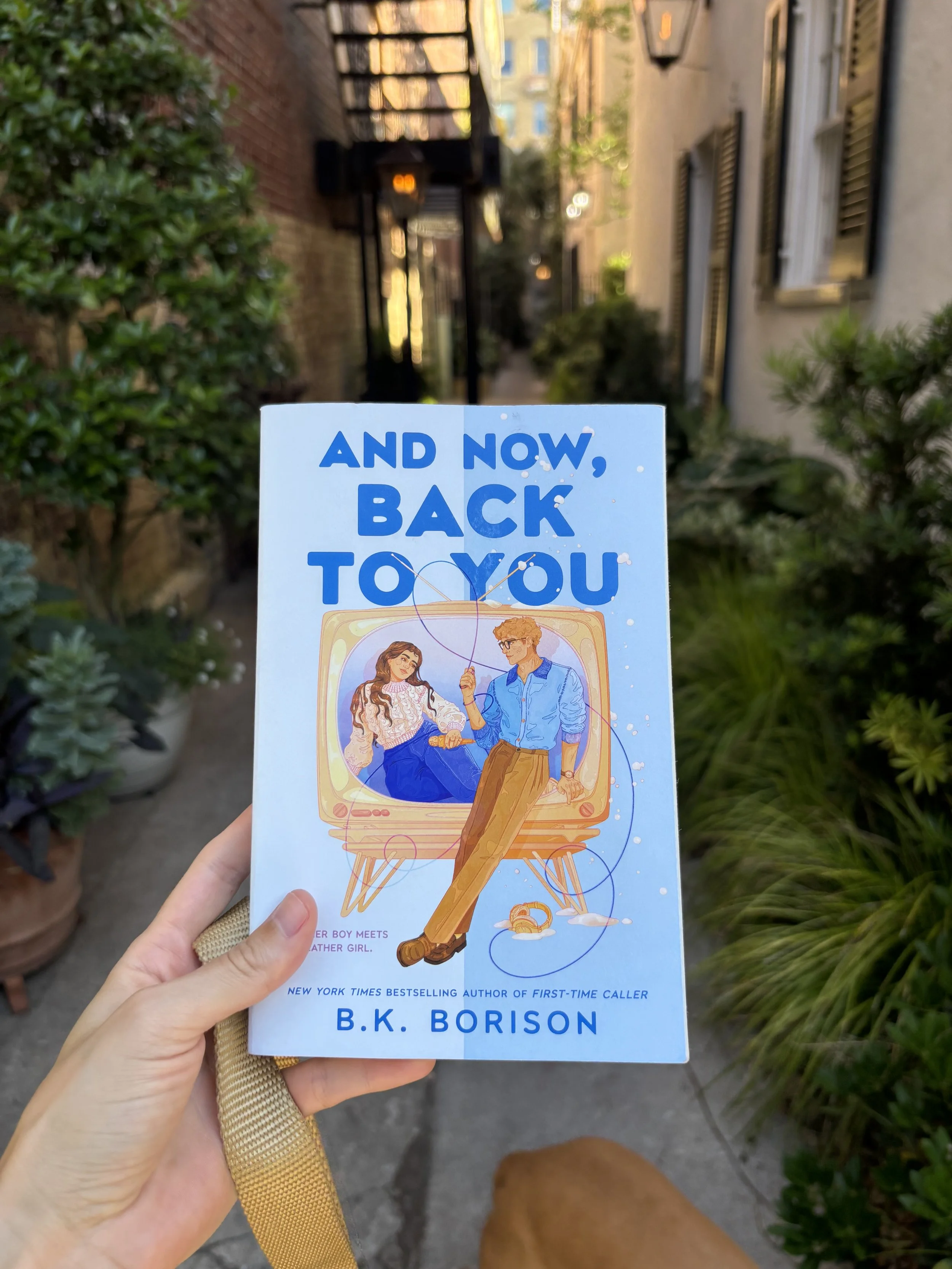 A person holding a book titled 'And Now, Back to You' by B.K. Borison outdoors on a city sidewalk with plants and apartment buildings in the background.