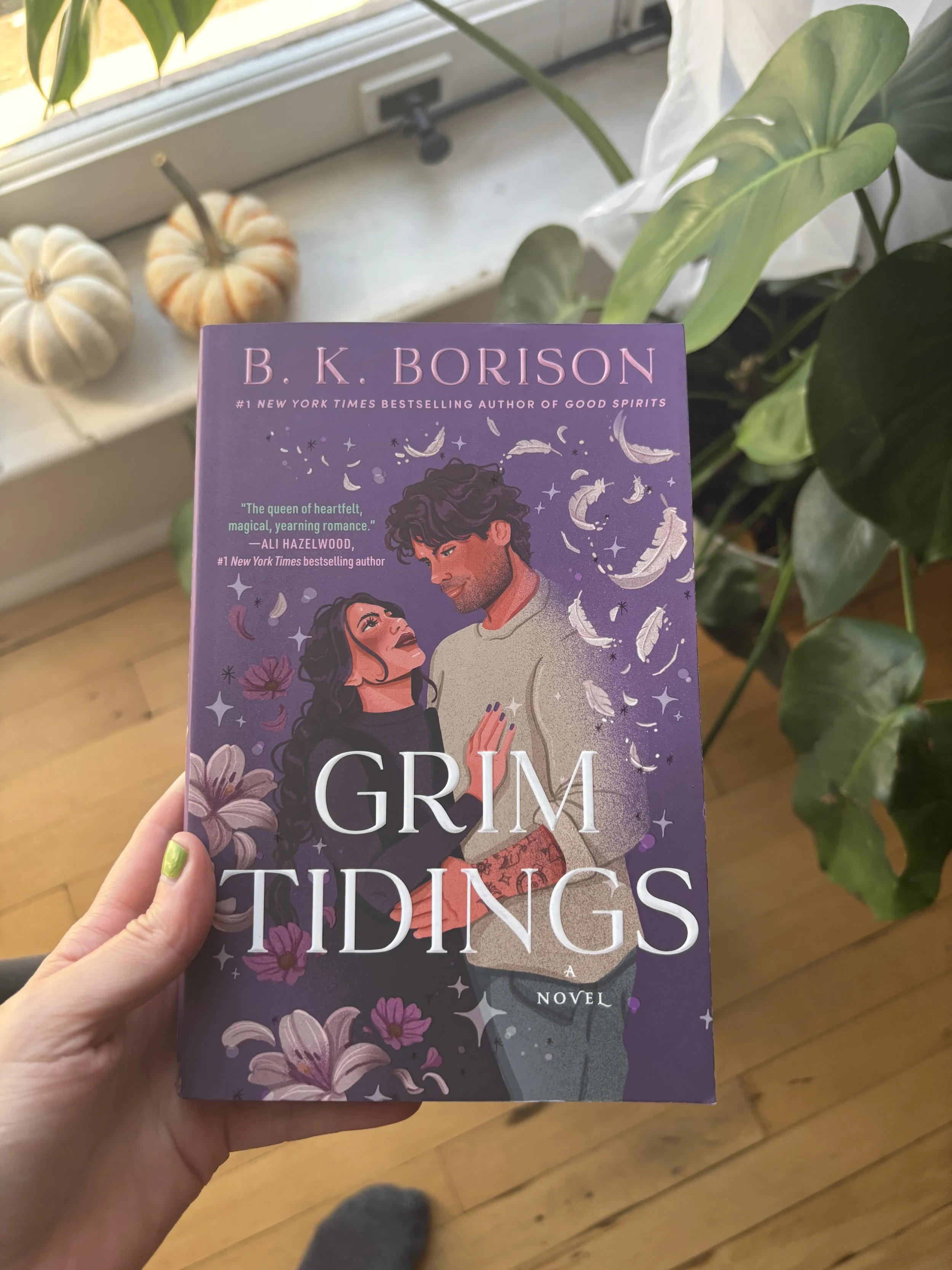 A person holding a purple paperback book titled "Grm Tidings" by B. K. Borison. The cover features an illustration of a man and woman looking at each other, surrounded by feathers and flowers. In the background, there are two small decorative pumpkins on a windowsill and a plant with large green leaves.