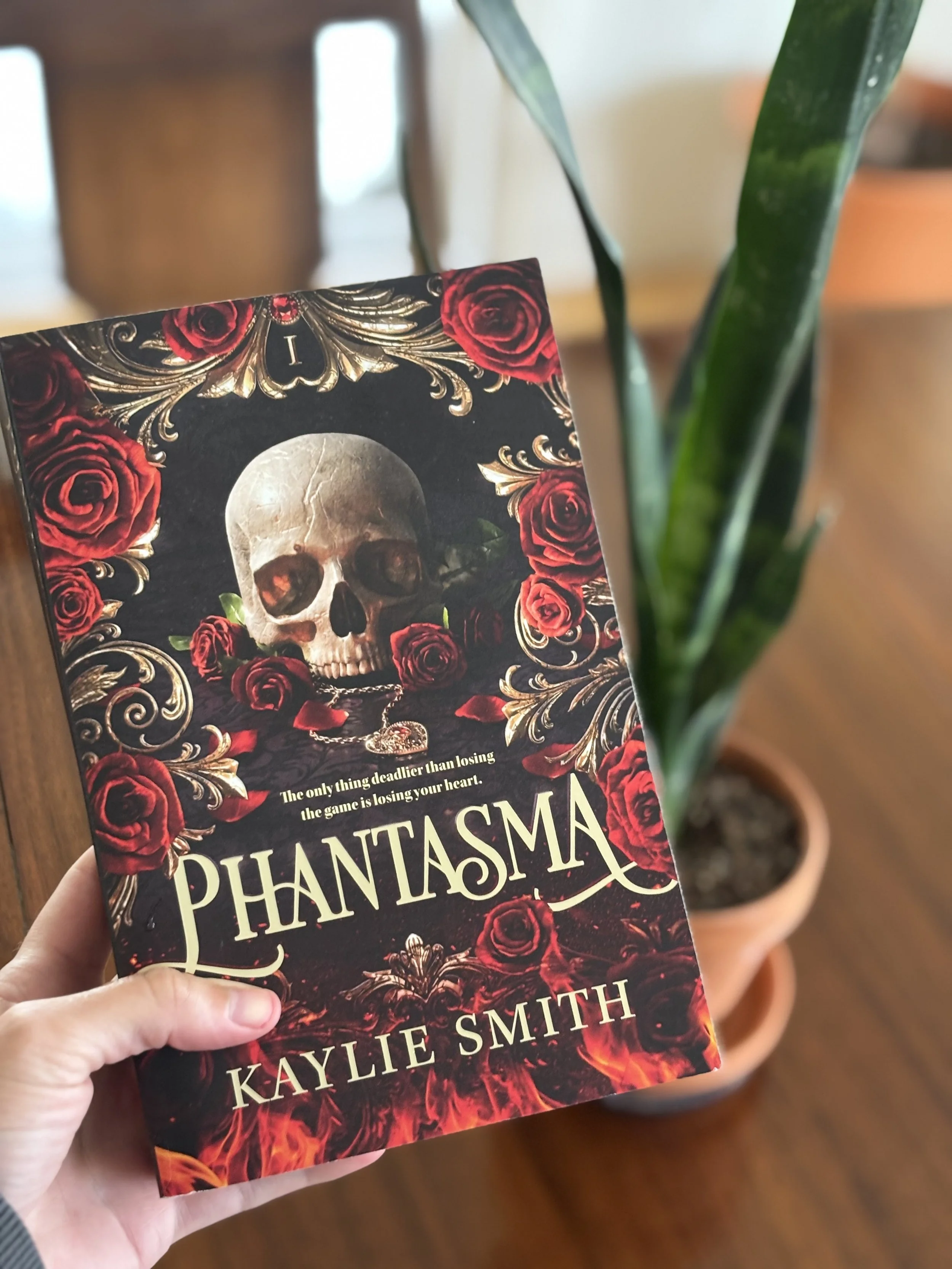 A person holding a book titled 'Phantasma' by Kaylie Smith, featuring a skull surrounded by red roses and intricate gold patterns on the cover.