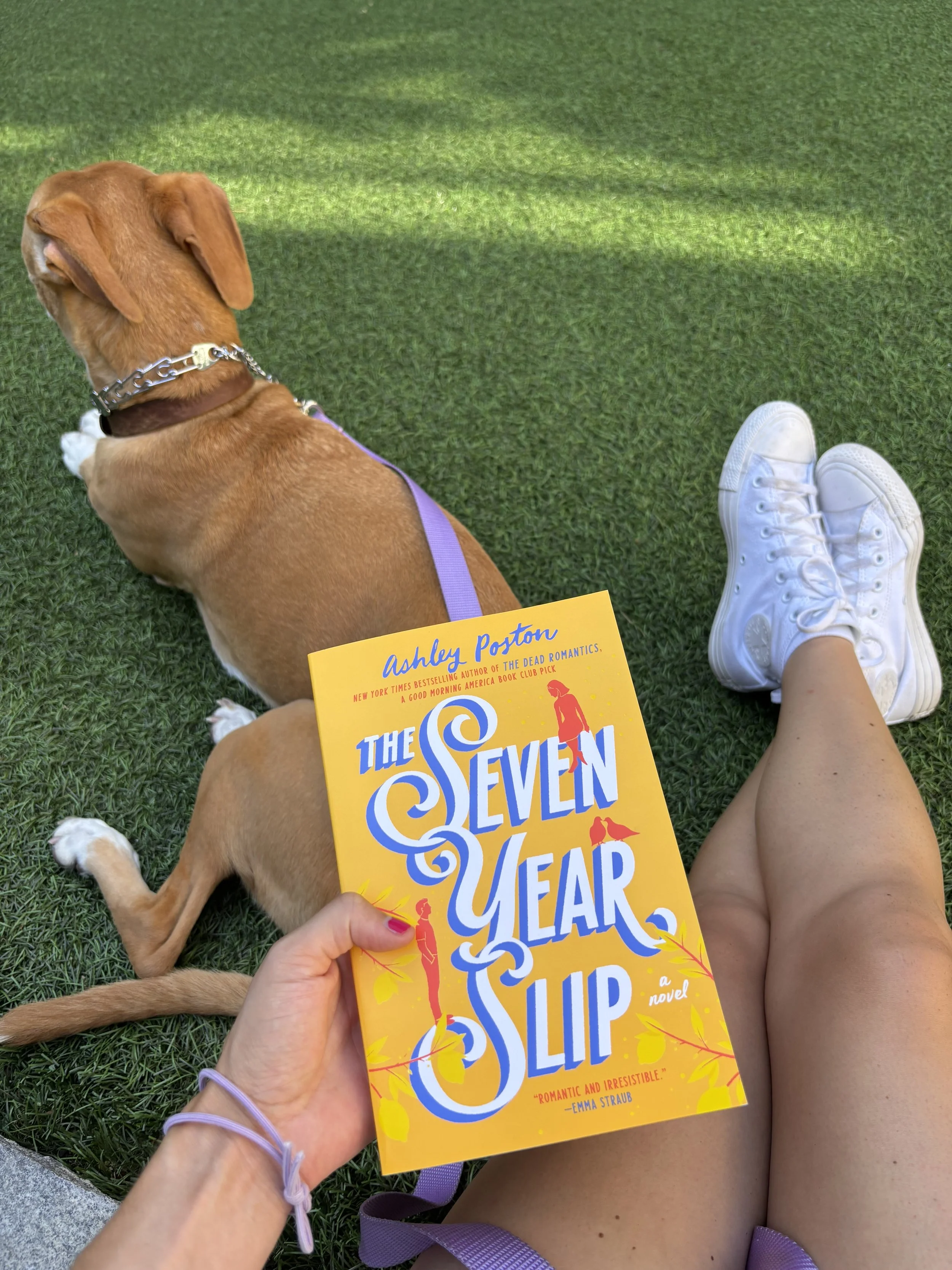 Person sitting on grass holding a bright yellow book titled 'The Seven Year Slip' with white sneakers visible, a tan dog lying beside them, and lush green grass in the background.