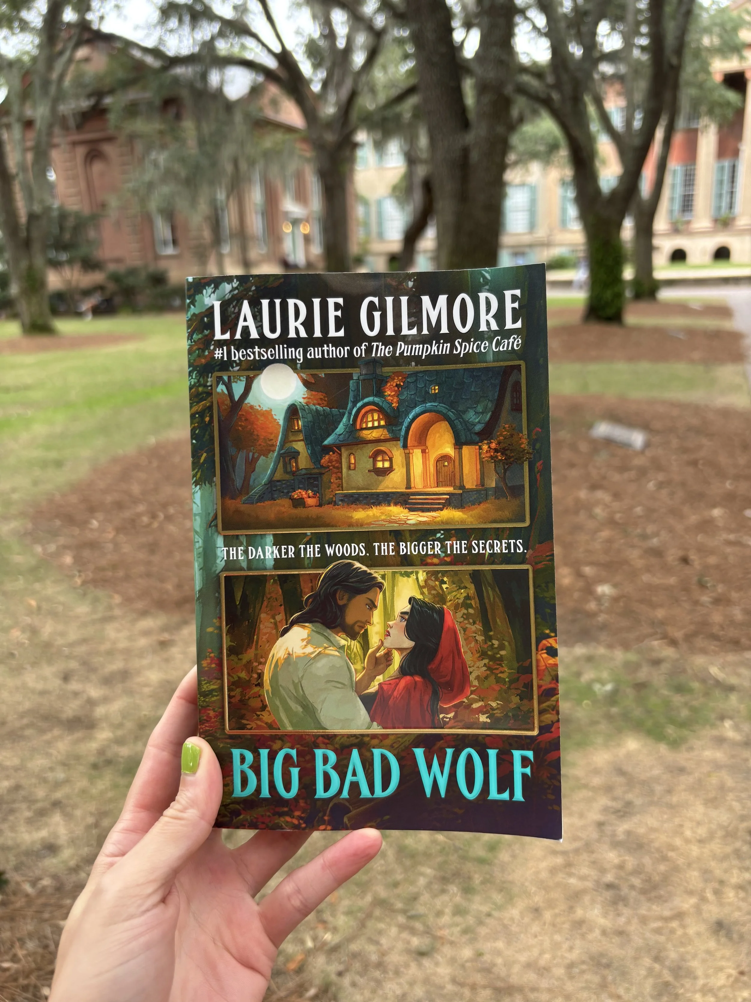 Close-up of a person holding a paperback book titled "Big Bad Wolf" by Laurie Gilmore outdoors with trees and historic buildings in the background.