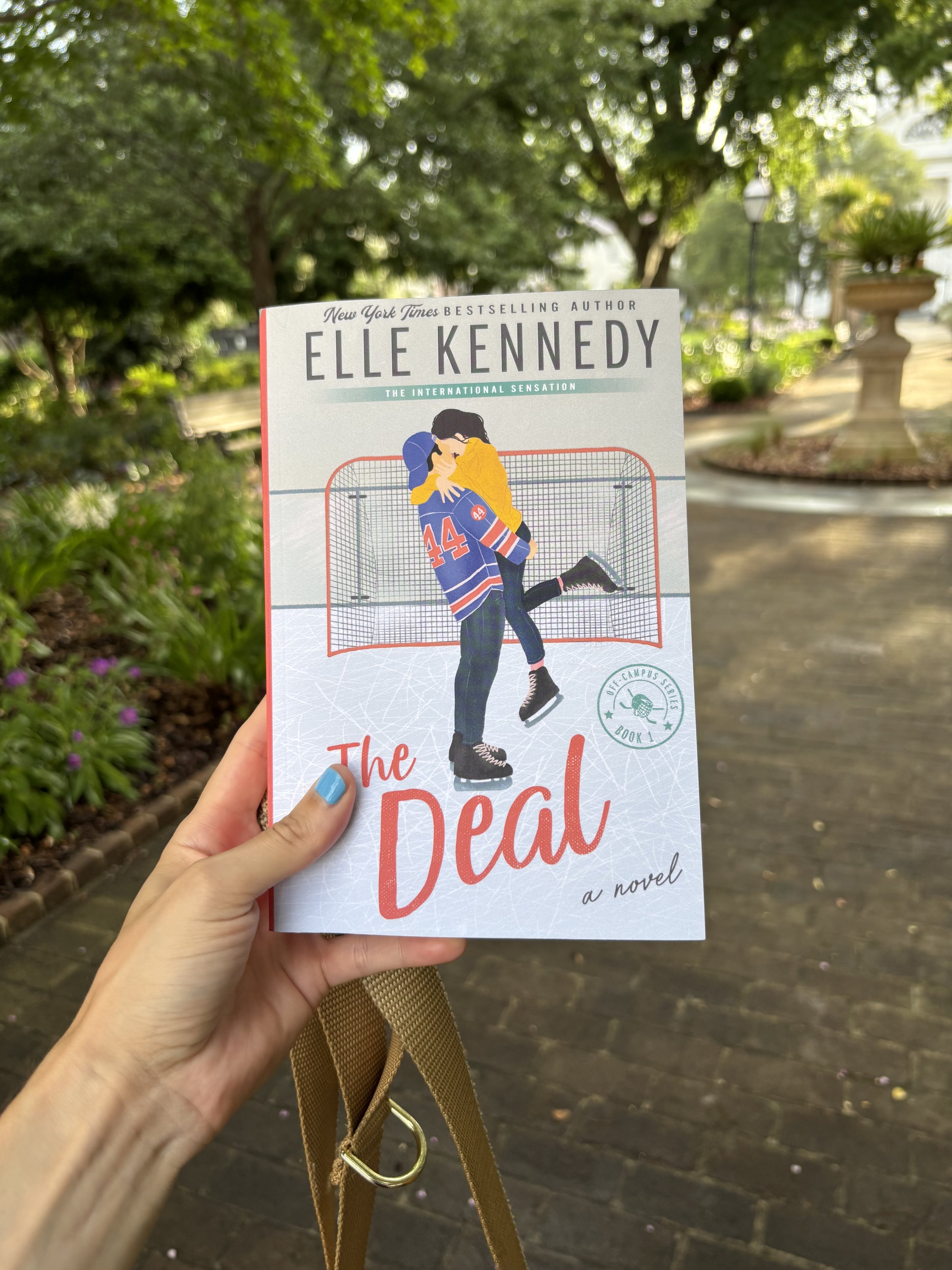 Book cover titled "The Deal" by Elle Kennedy, held in a person's hand outdoors with trees and a park path in the background.