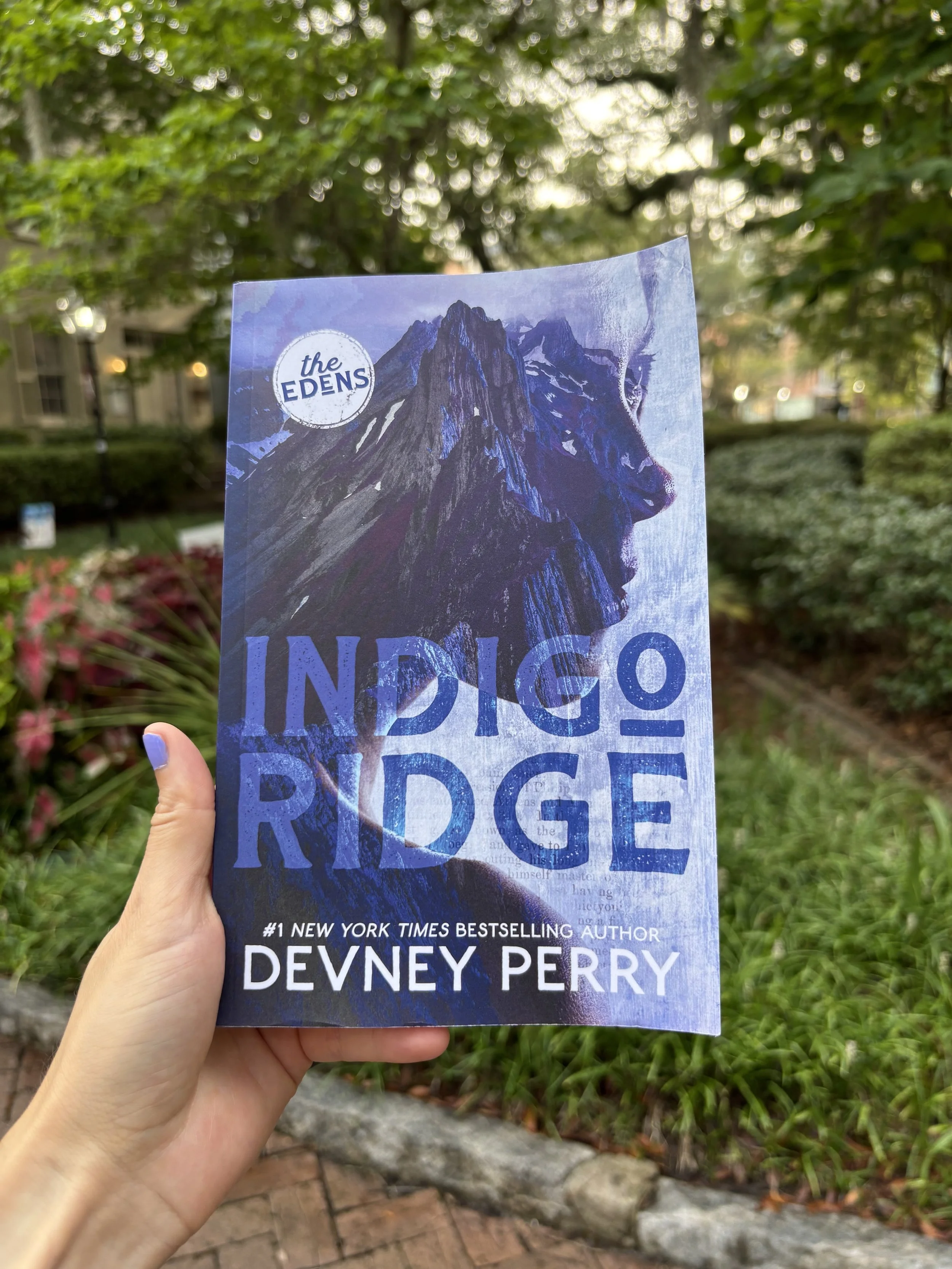 A hand holding a copy of the book 'Indigo Ridge' by Devney Perry outdoors with trees and bushes in the background.