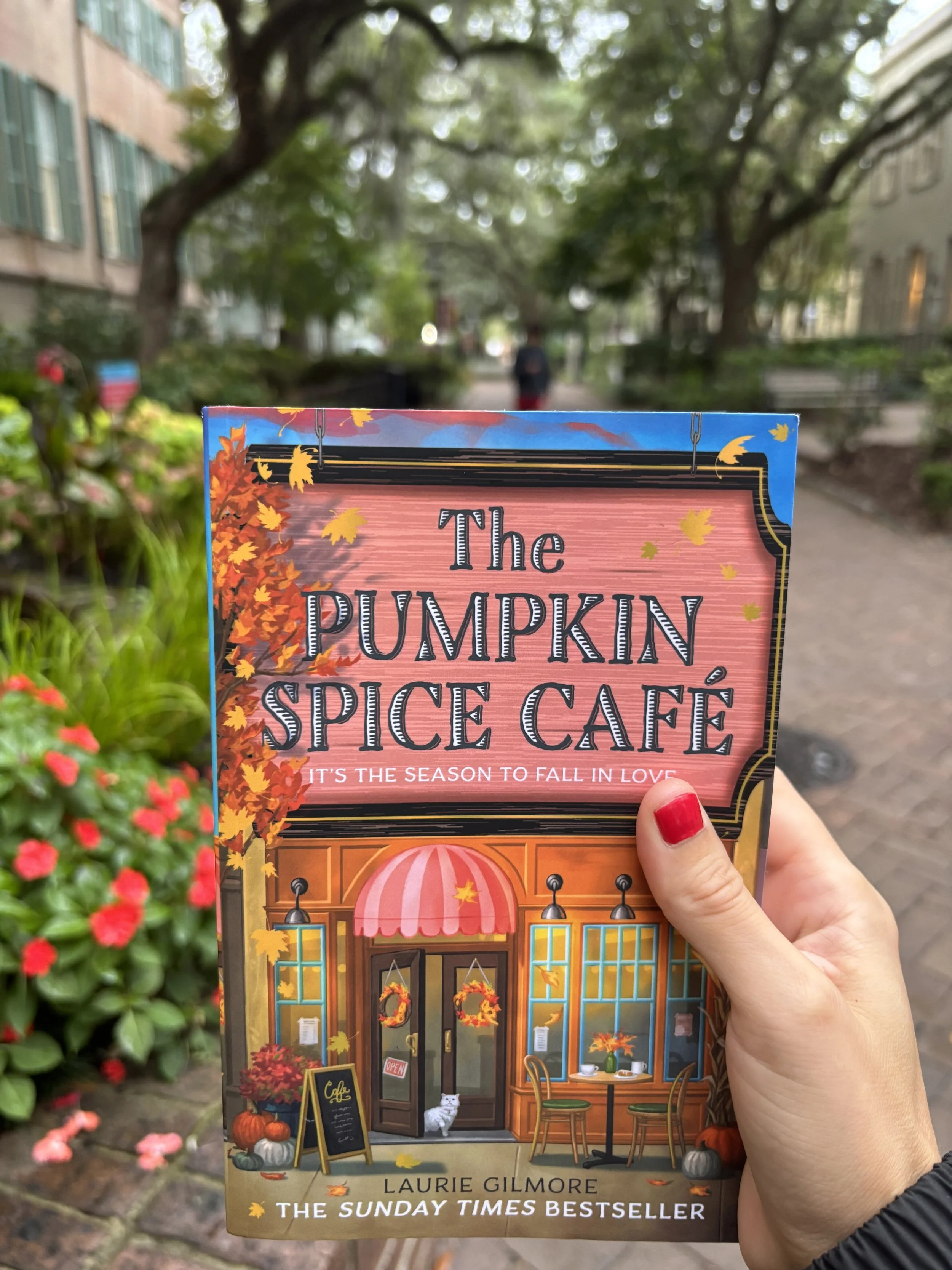 A person holding a book titled "The Pumpkin Spice Café" outdoors with trees and buildings in the background. The book cover shows a cozy café scene with pumpkins and fall decorations.