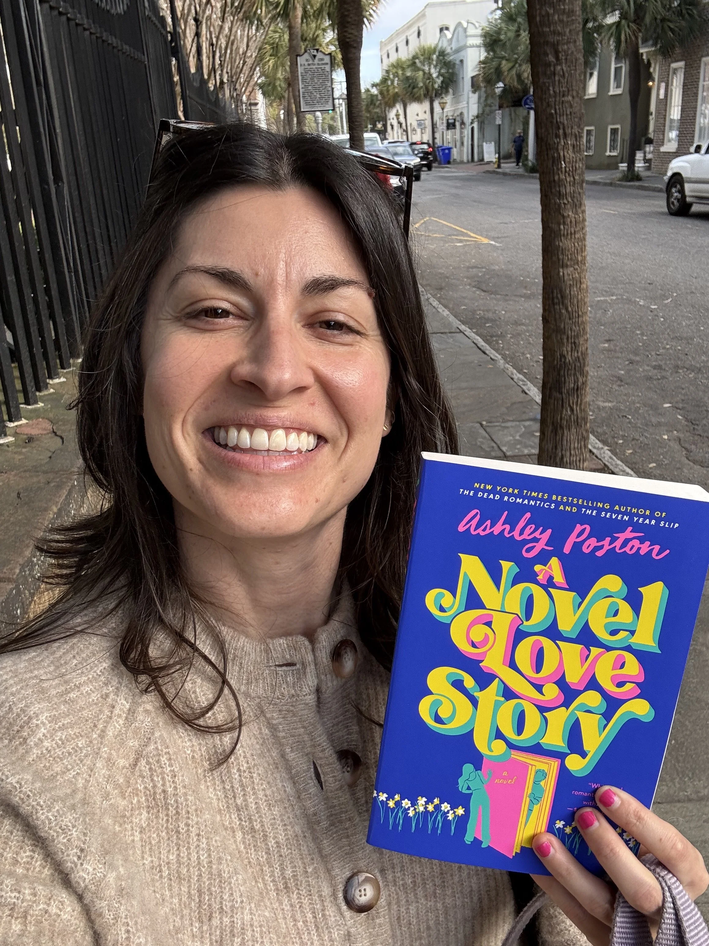 A woman smiling while holding a colorful book titled 'A Novel Love Story' by Ashley Poston outside on a city sidewalk lined with trees, buildings, and parked cars.