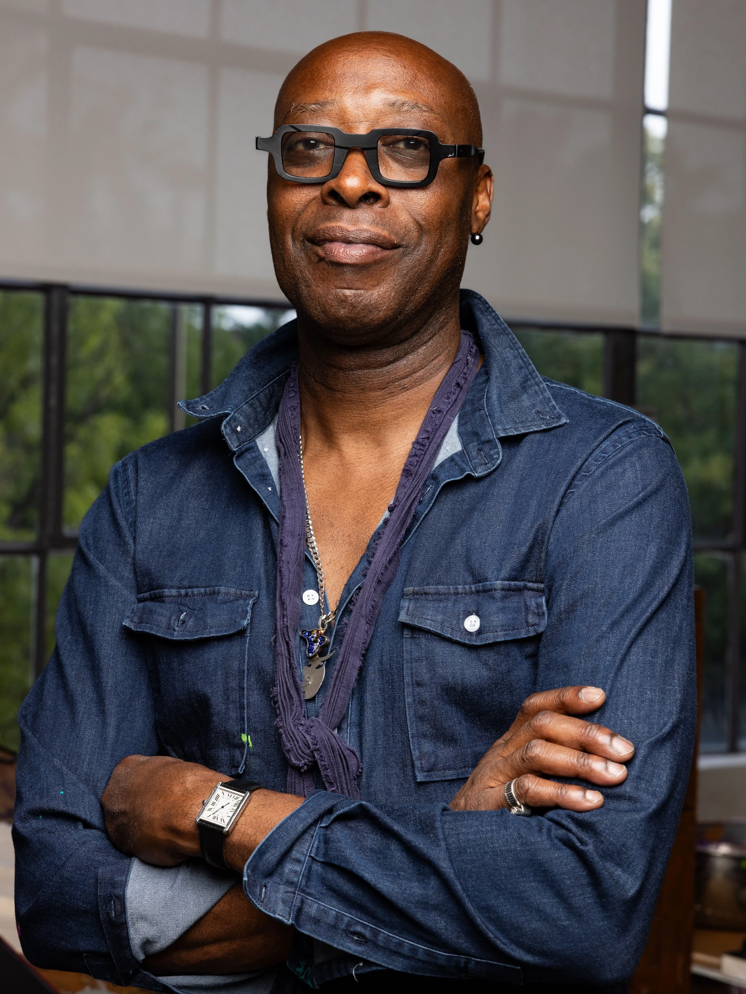 A confident middle-aged man with glasses and a bald head, wearing a denim shirt with rolled-up sleeves, standing indoors in front of large windows with greenery outside. He has his arms crossed and is looking at the camera with a slight smile.