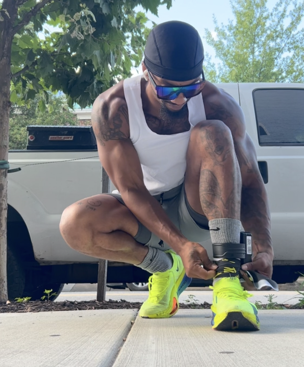 A man in athletic gear, sunglasses, and a black durag, tying his bright yellow running shoes outdoors next to a white truck.