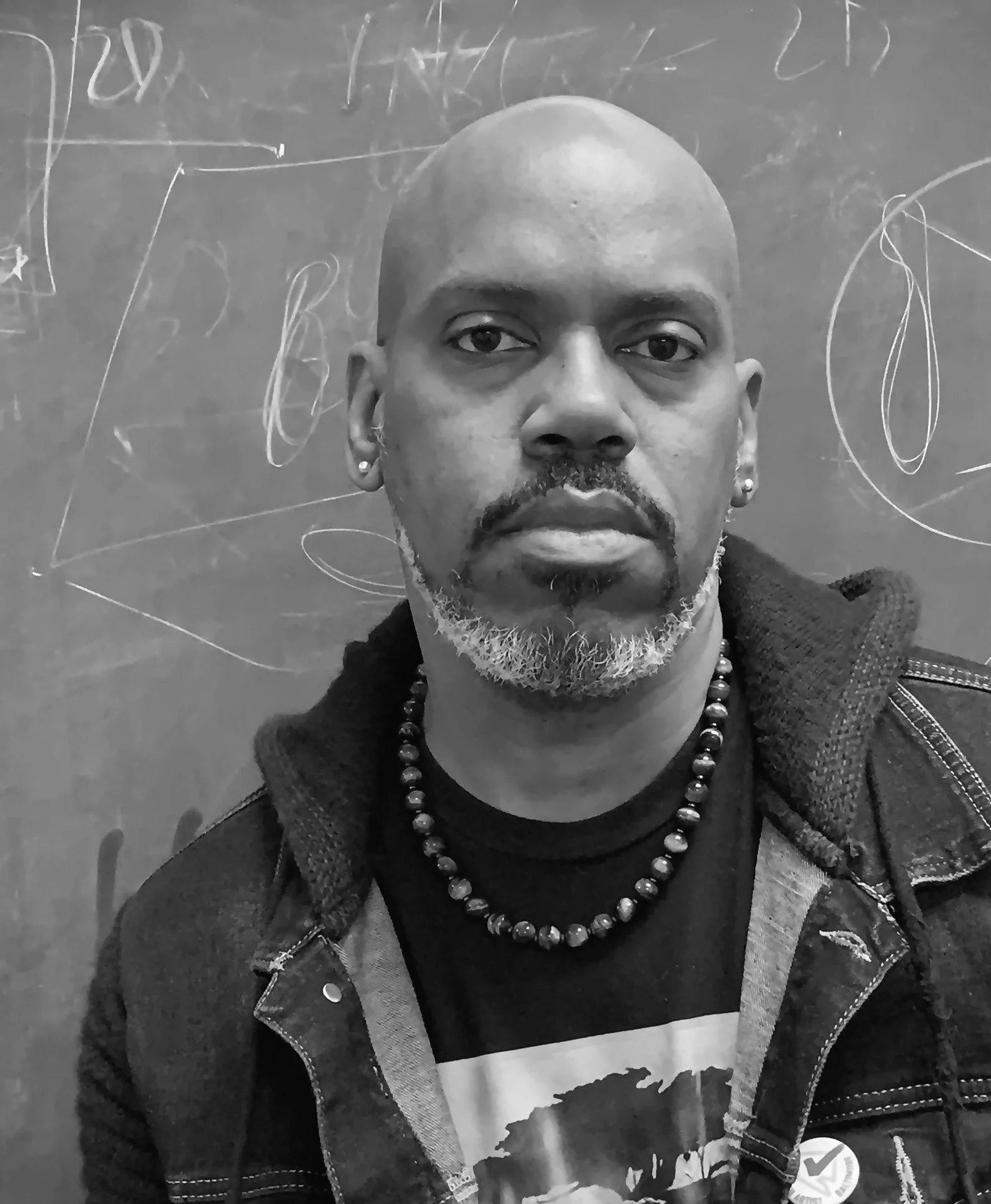 A black-and-white photo of a man with a bald head, a gray beard, and earrings. He's wearing a beaded necklace, a printed t-shirt, and a denim jacket, standing in front of a chalkboard with writing and drawings.