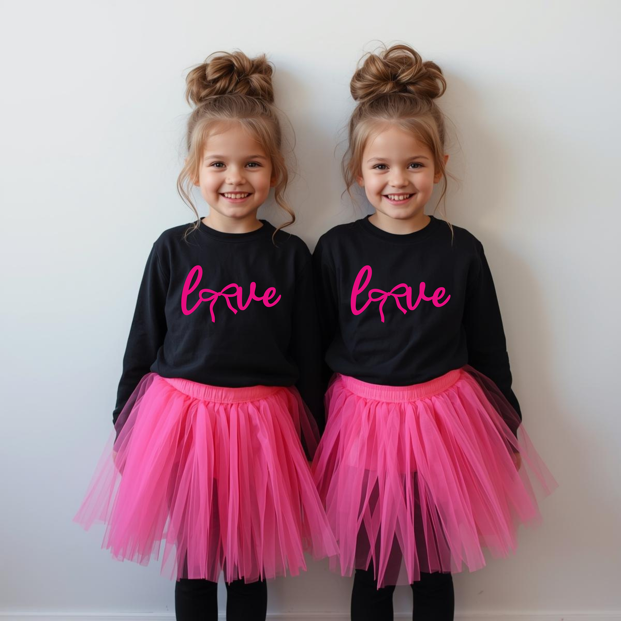 Twin girls wearing black shirts with pink 'love' text and pink tutu skirts, standing against a white wall, smiling.