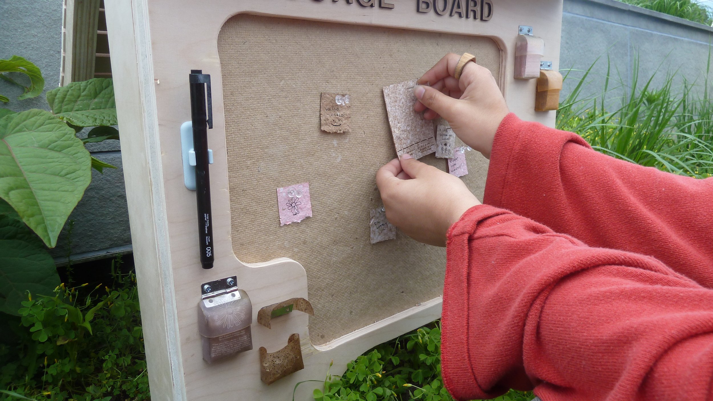 A person with a red long-sleeve shirt pins small pieces of paper with drawings and writing onto a cork bulletin board outdoors, surrounded by green plants.
