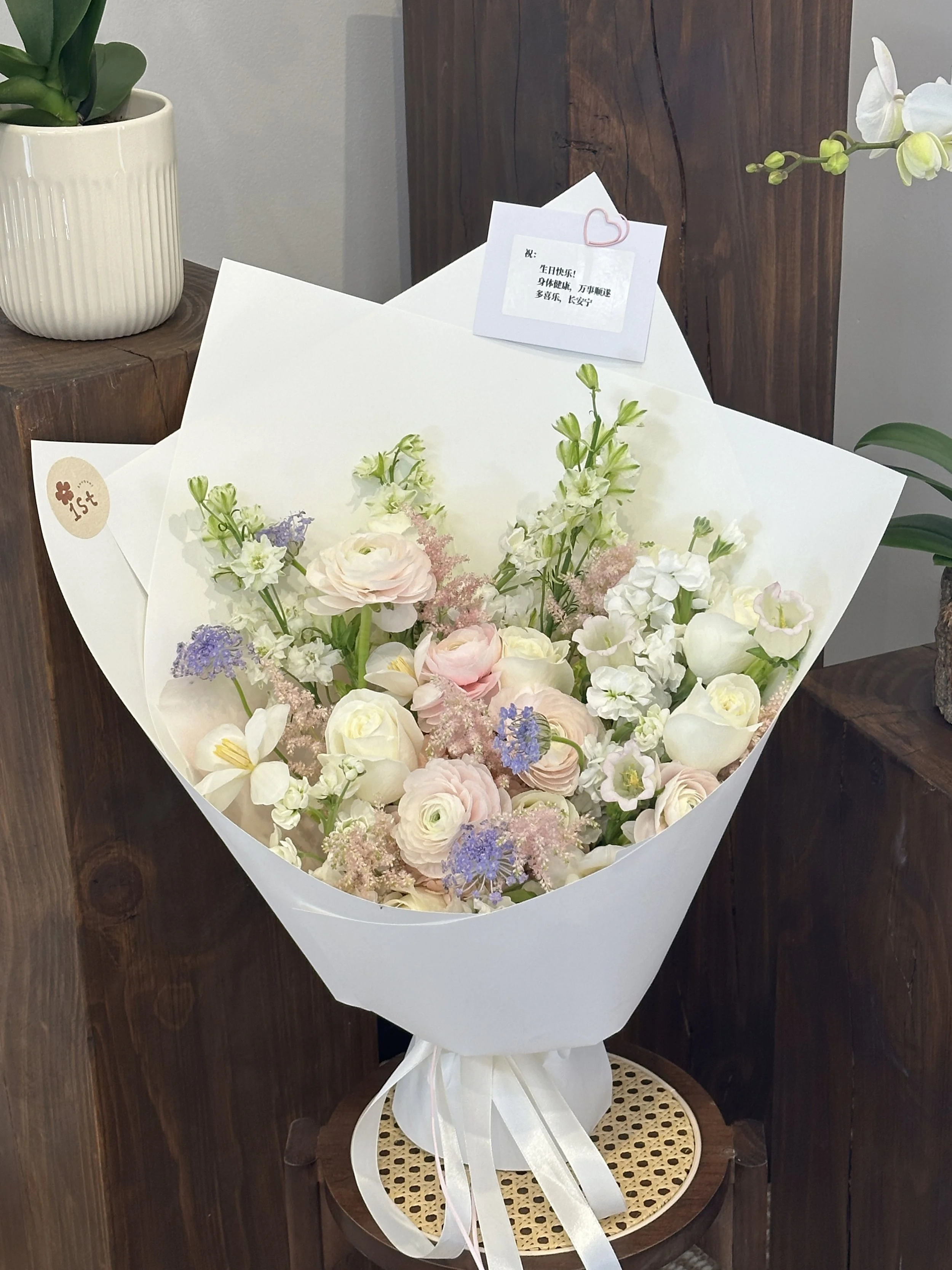 A bouquet of pastel-colored roses, ranunculus, and other flowers. It is wrapped in white paper with a ribbon at the bottom, placed on a small wooden stool. There is a note with pink heart paper clip and a small cream-colored card attached to the bouquet. The background includes a wooden plank and potted plants.