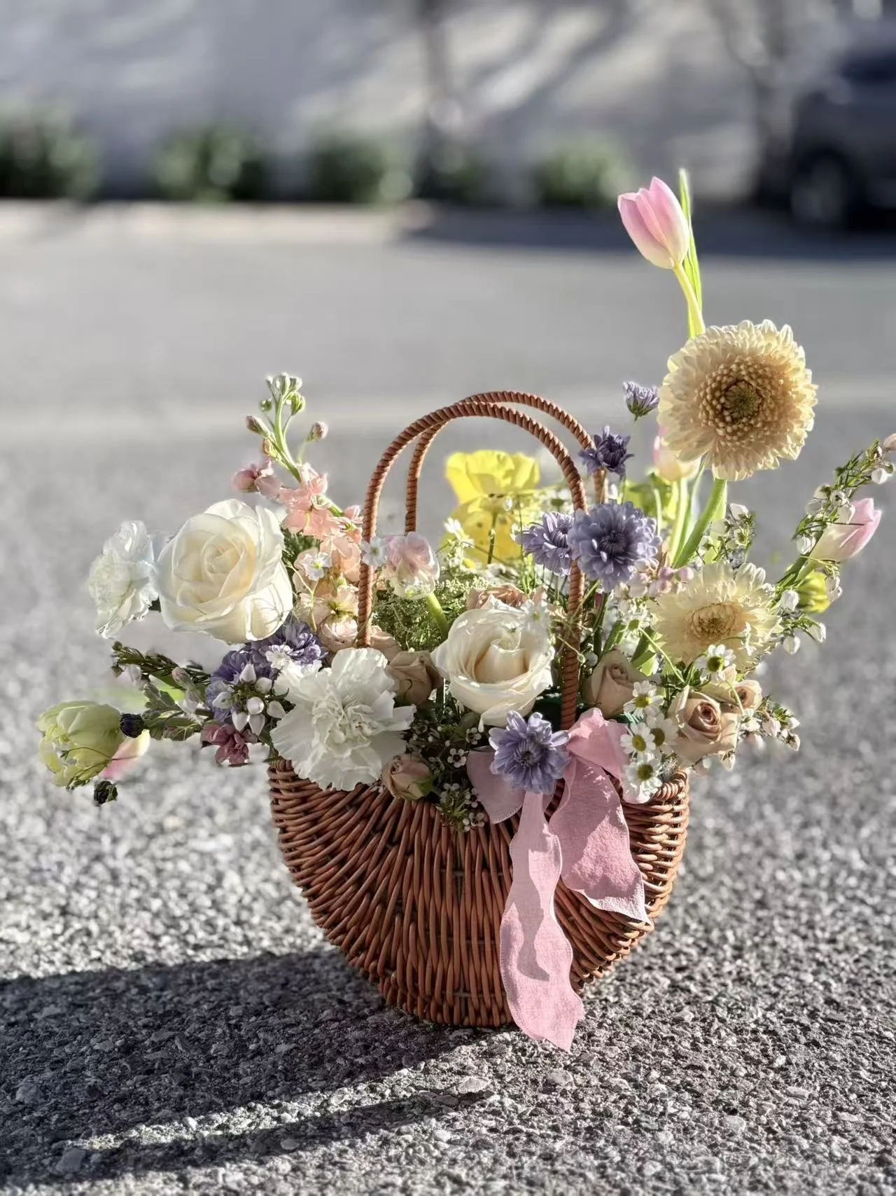 A woven basket filled with assorted pastel-colored flowers, including roses, daisies, and tulips, placed on a textured surface outdoors.