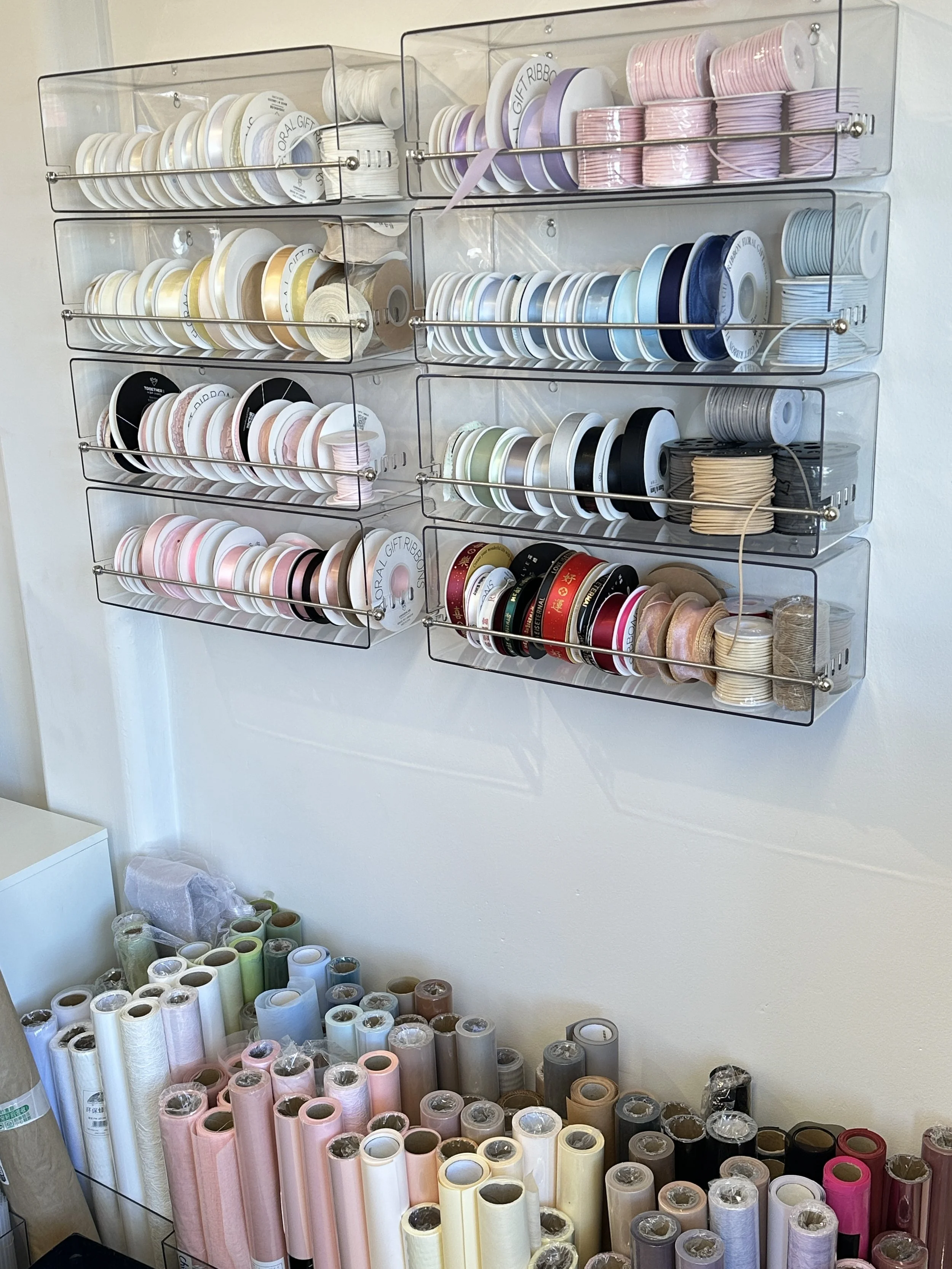 A wall-mounted organization system with clear plastic compartments holding spools of ribbon in various colors and patterns, with loose spools of ribbon stored in boxes below.