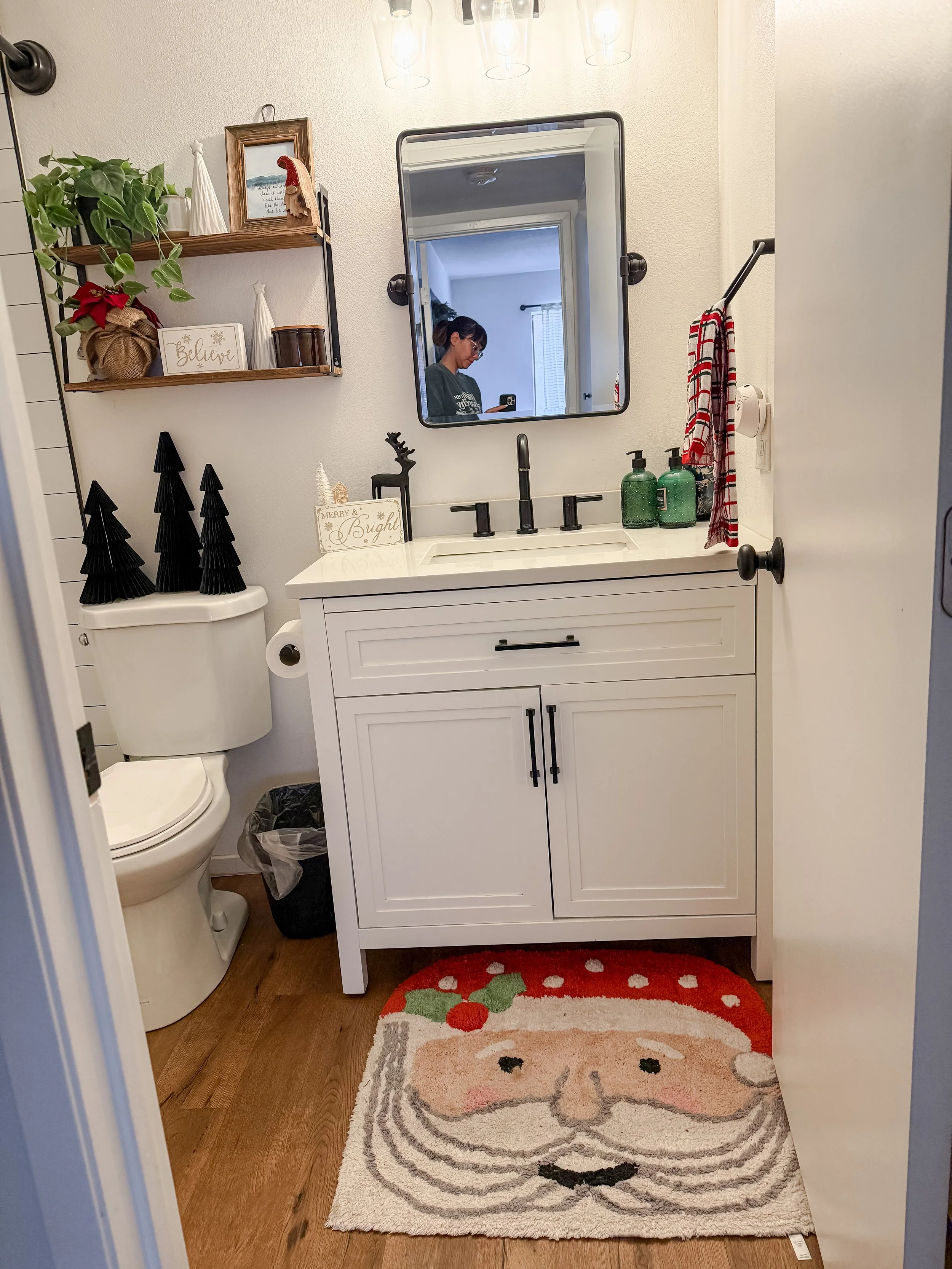 A small bathroom decorated for Christmas with a Santa face rug, black Christmas trees on the toilet tank, shelves with holiday decorations, a mirror, and a person taking a selfie.