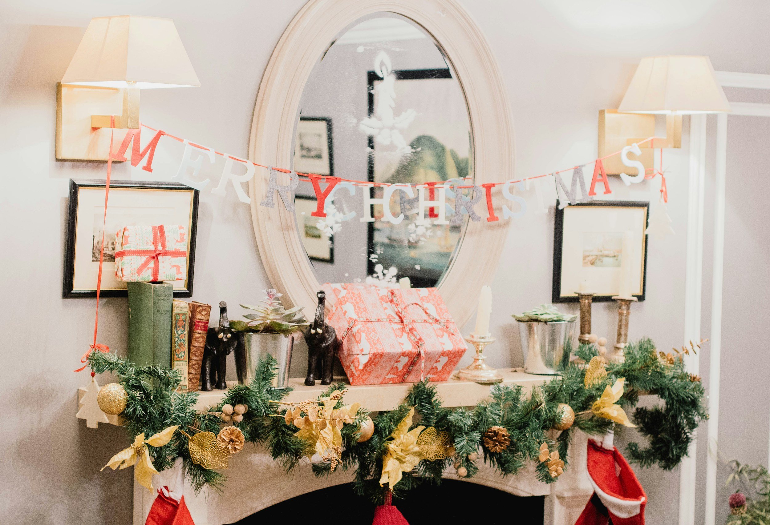 A decorated fireplace mantel with Christmas decorations, including a 'Merry Christmas' banner, wrapped presents, greenery, ornaments, candles, and stockings.