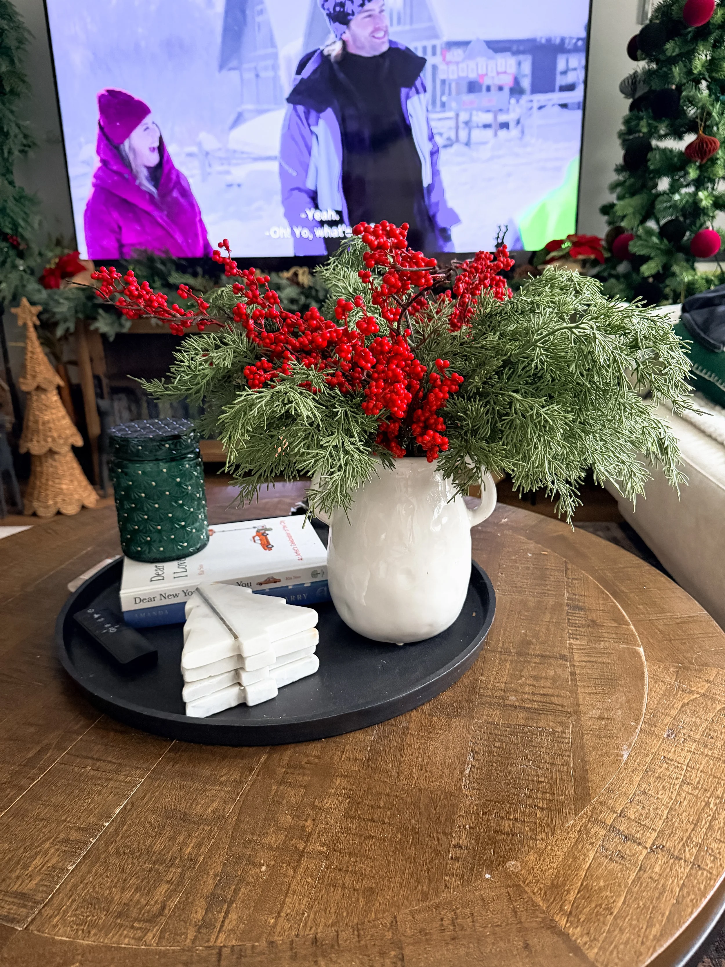 A white vase with red berries and green foliage on a black tray on a wooden table, with books, a white stack, and a remote, in a room decorated for Christmas, with a TV showing two people in winter clothing and Christmas trees in the background.
