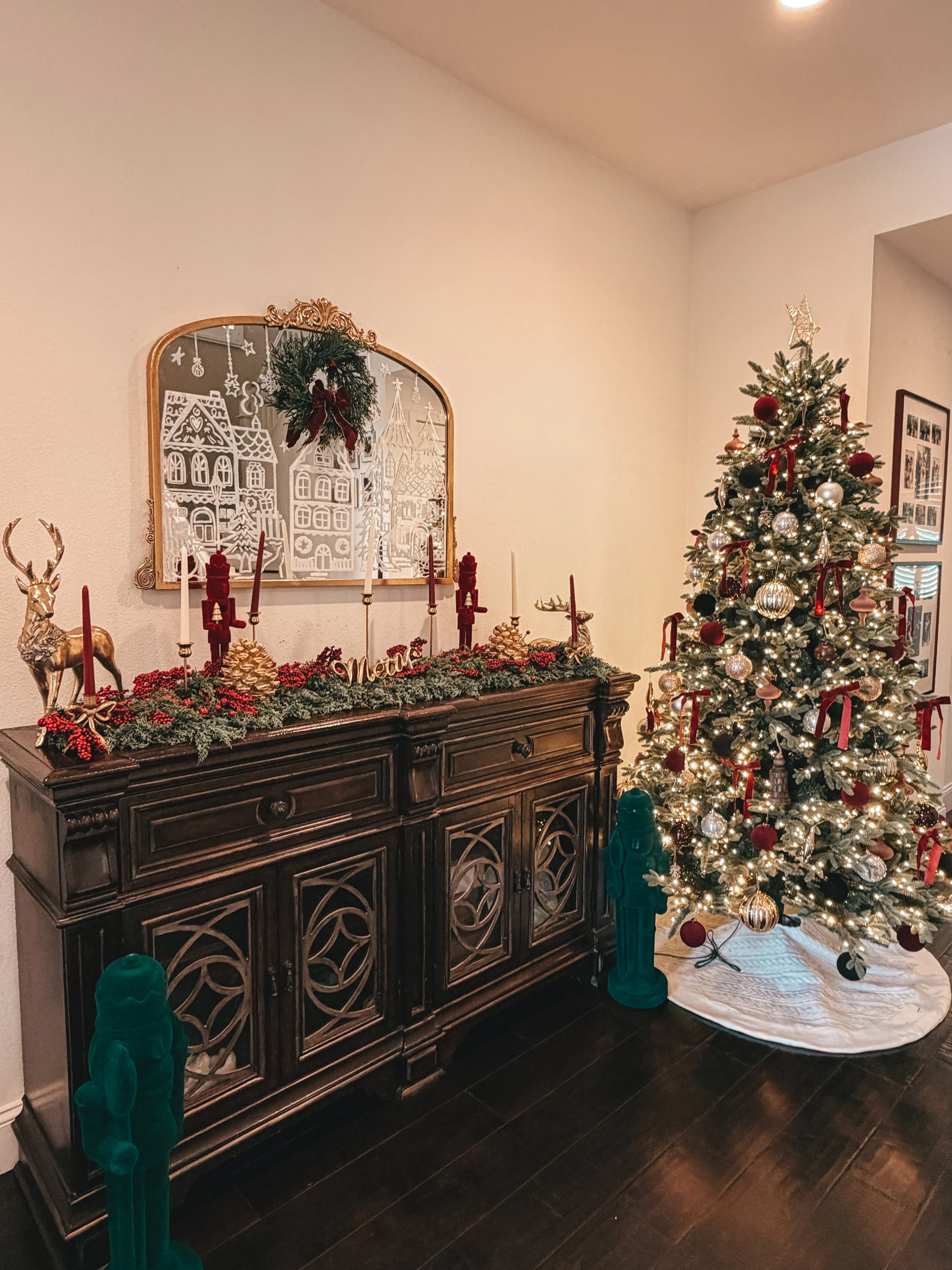 A decorated Christmas tree with ornaments and lights, positioned next to a dark wooden sideboard decorated with candle holders, pinecones, and a 'Merry' sign, with framed photos on the wall behind.