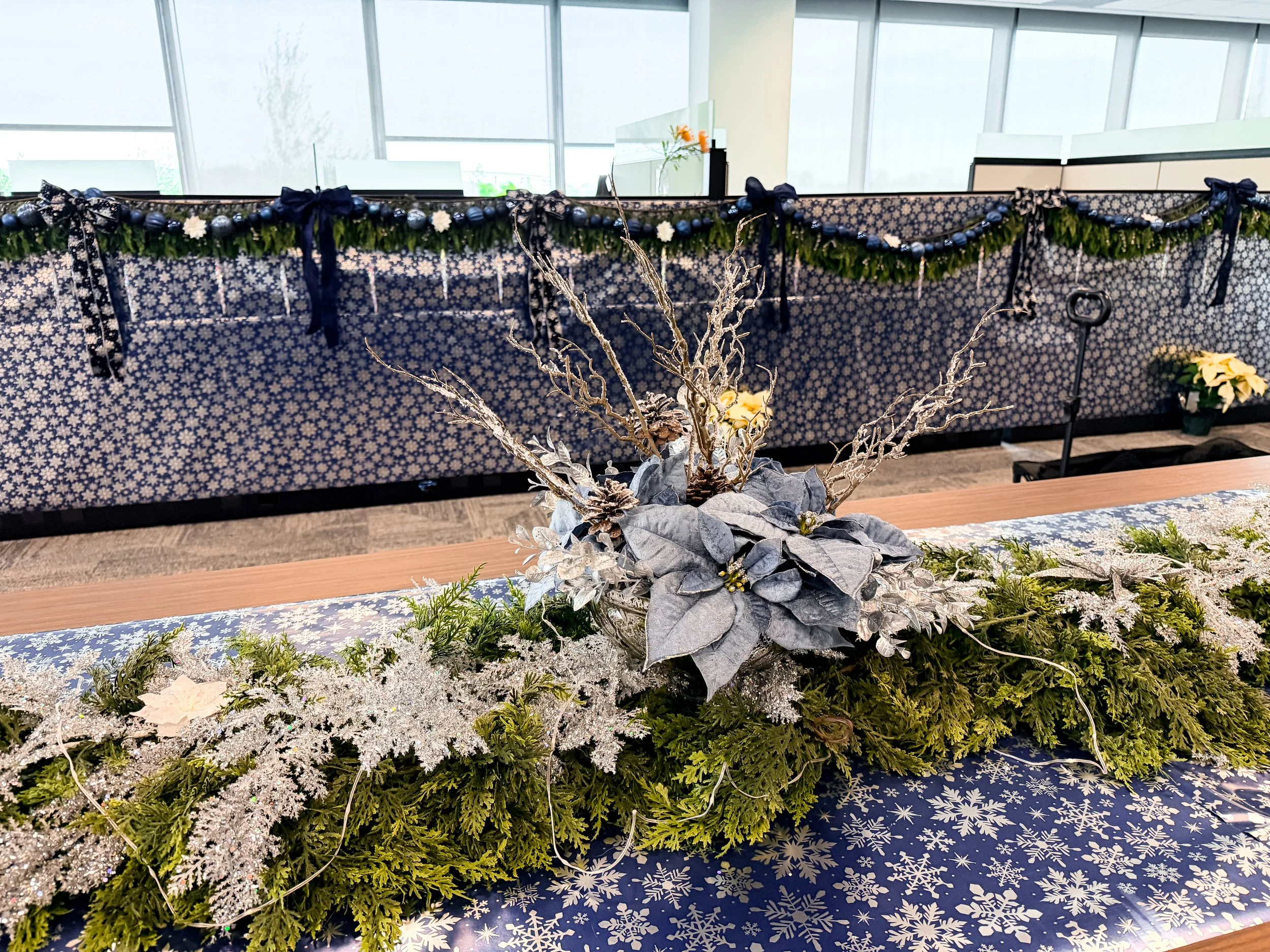 Christmas table centerpiece with silver and green decorations, including leaves and branches, on a table with snowflake-patterned tablecloth. In the background, a decorated partition wall with garland, bows, and ornaments.