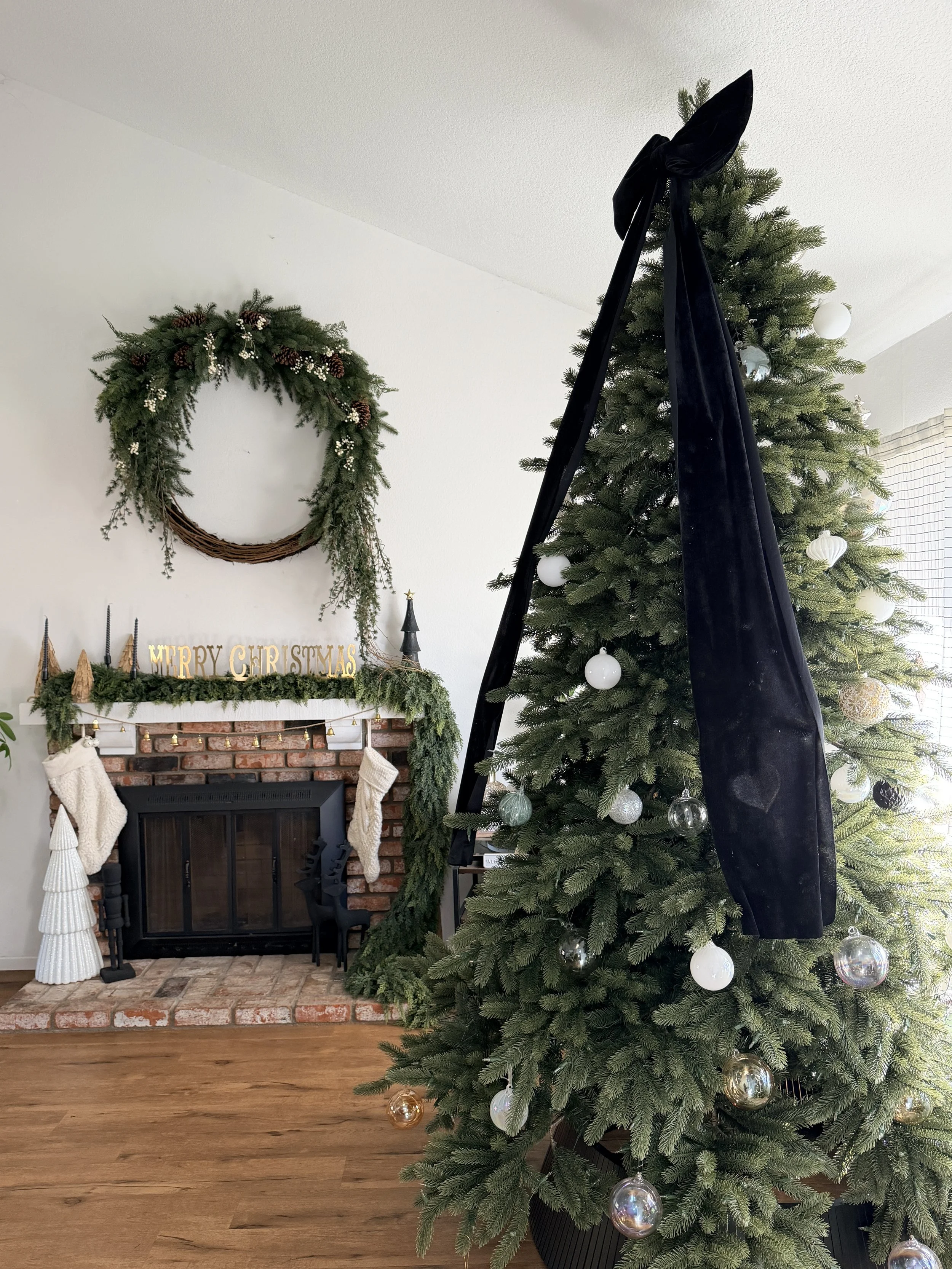 A decorated Christmas tree positioned next to a brick fireplace with stockings hanging, a green garland on the mantel, and a wreath hanging on the wall above. The room has wooden flooring and a window with blinds letting in natural light.