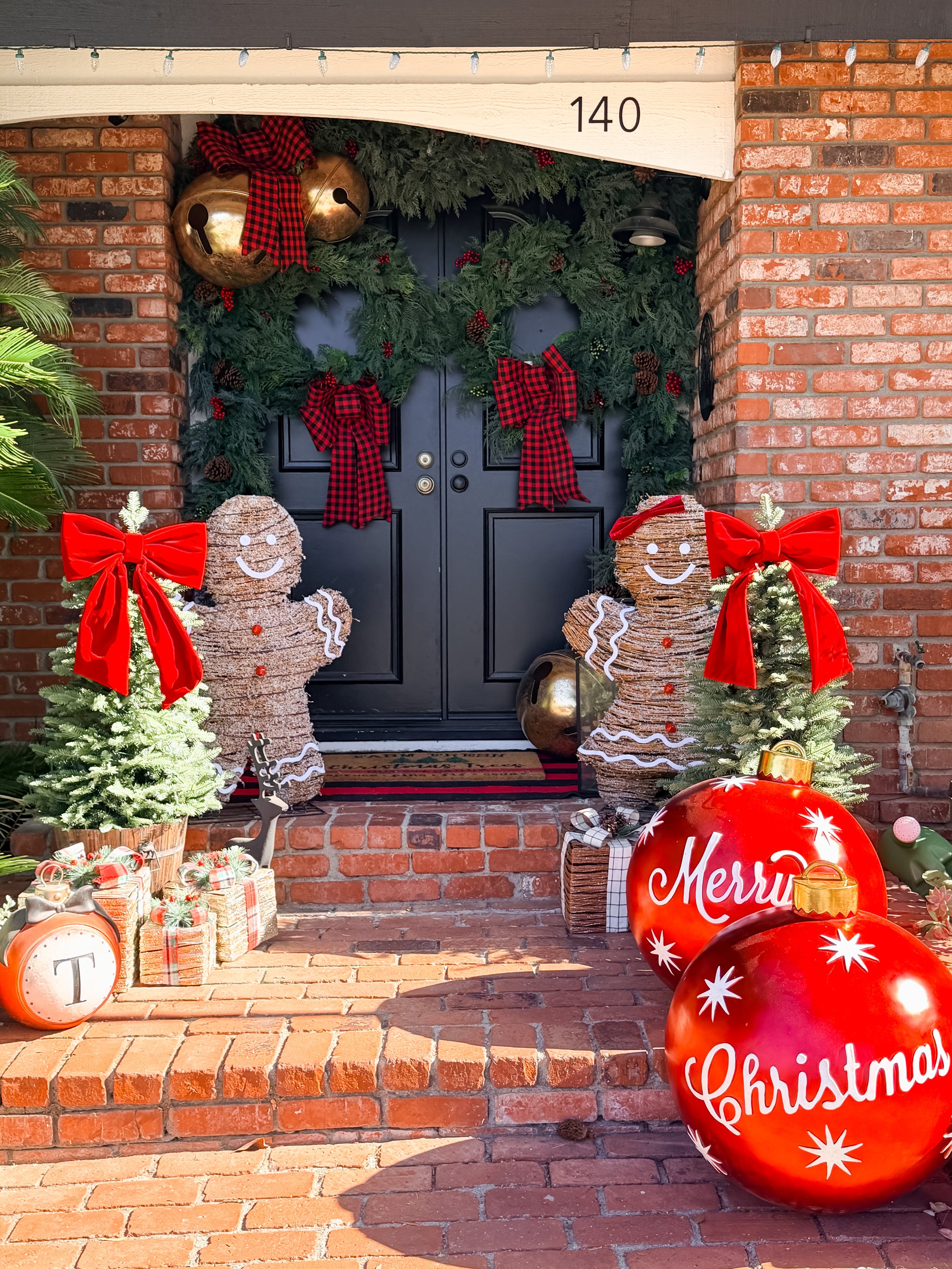 Decorated Christmas front porch with black door, greenery wreath with red plaid ribbons, gingerbread figure, small potted Christmas trees with large red bows, wrapped presents, and oversized red Christmas ornaments.