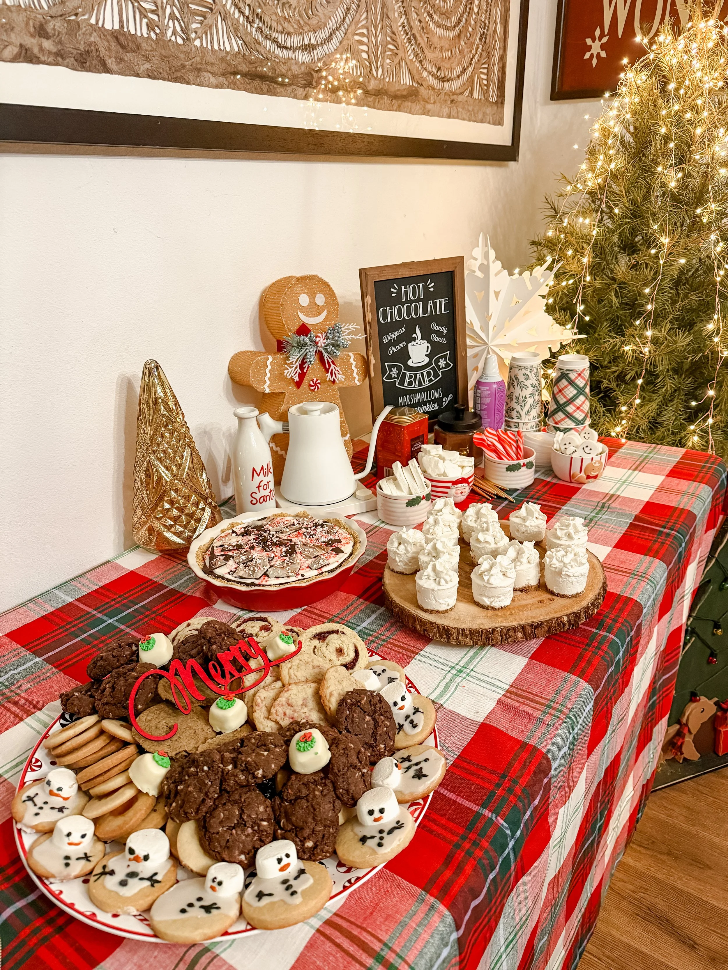 Christmas dessert table with cookies decorated as snowmen, chocolate chip cookies, a pie, marshmallows, and whipped cream decorated for the holidays. A small chalkboard sign reads 'Hot Chocolate.' A gingerbread man decoration and a Christmas tree wit